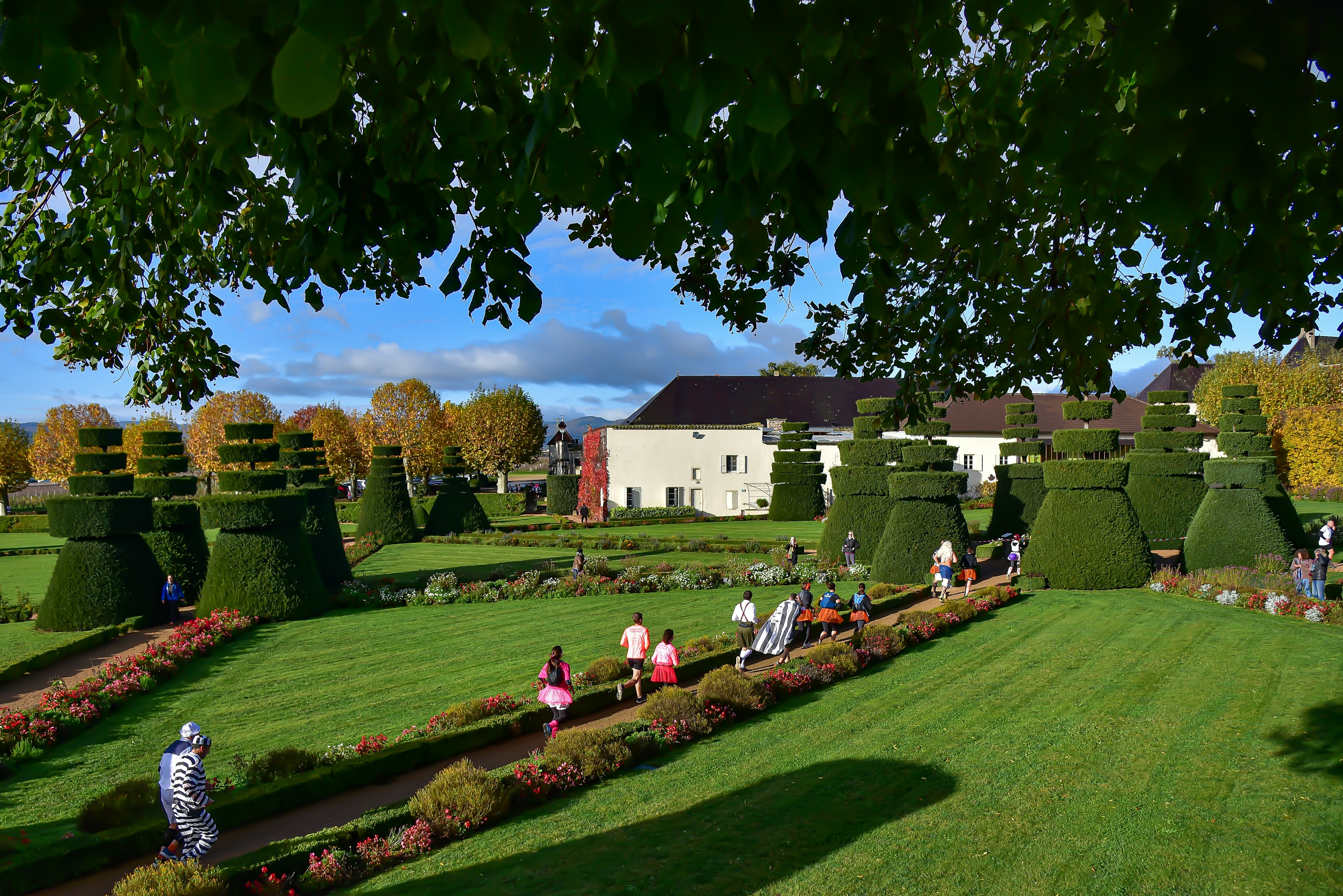 People wearing fancy dress costumes are running on a path through landscaped gardens with a manor house in the background