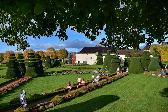 People wearing fancy dress costumes are running on a path through landscaped gardens with a manor house in the background