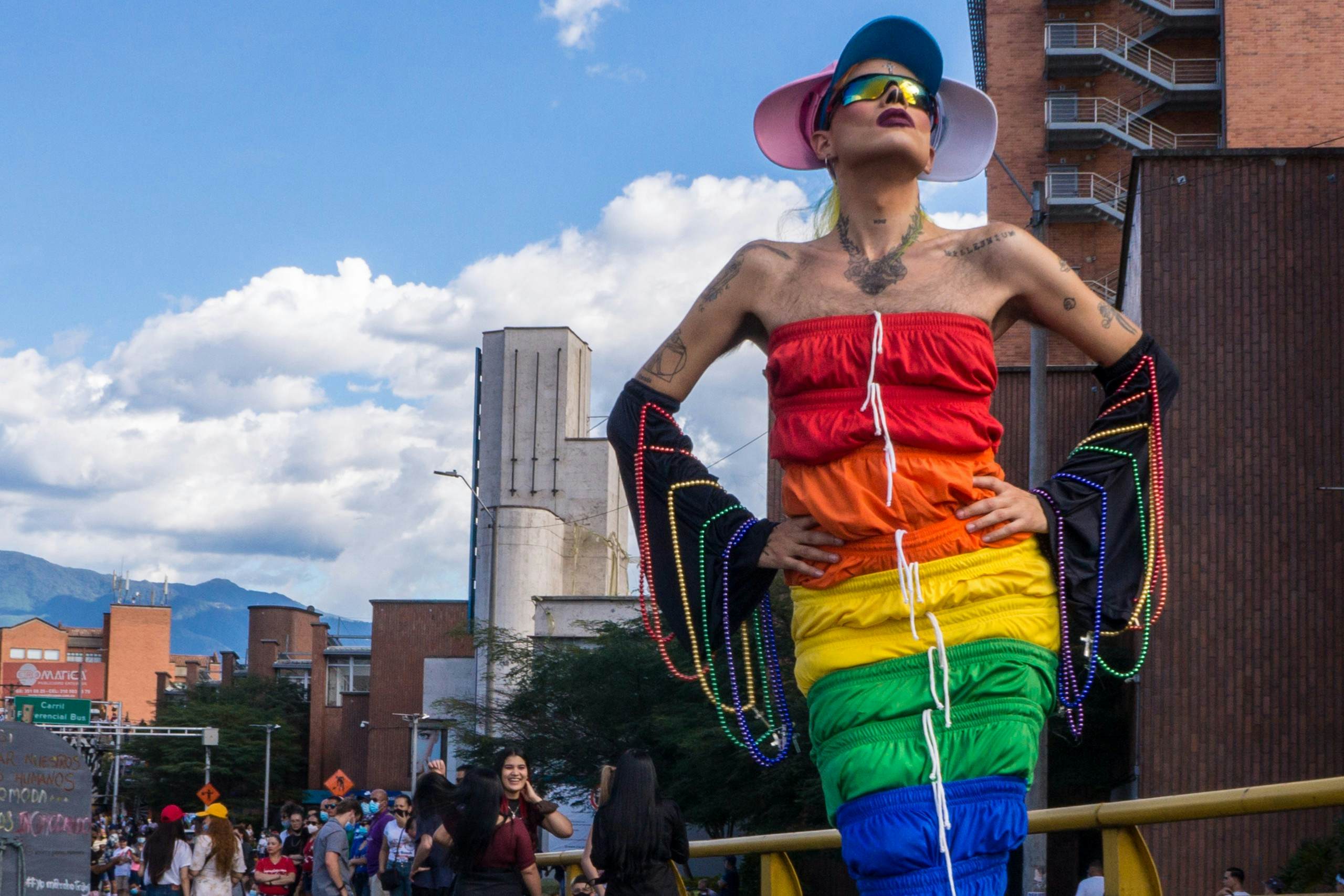 A drag queen dressed in a colorful rainbow-patterned dress draped with beads strikes a pose at an outdoor Pride event in a city