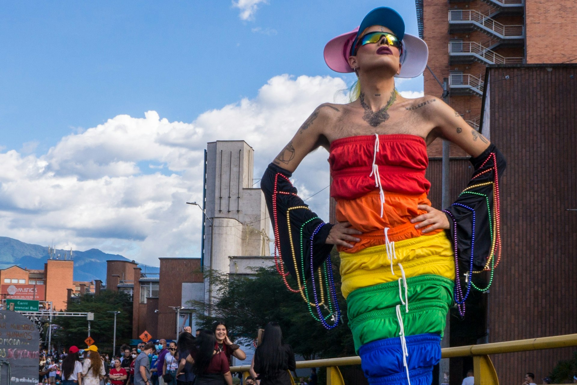 A drag queen dressed in a colorful rainbow-patterned dress draped with beads strikes a pose at an outdoor Pride event 