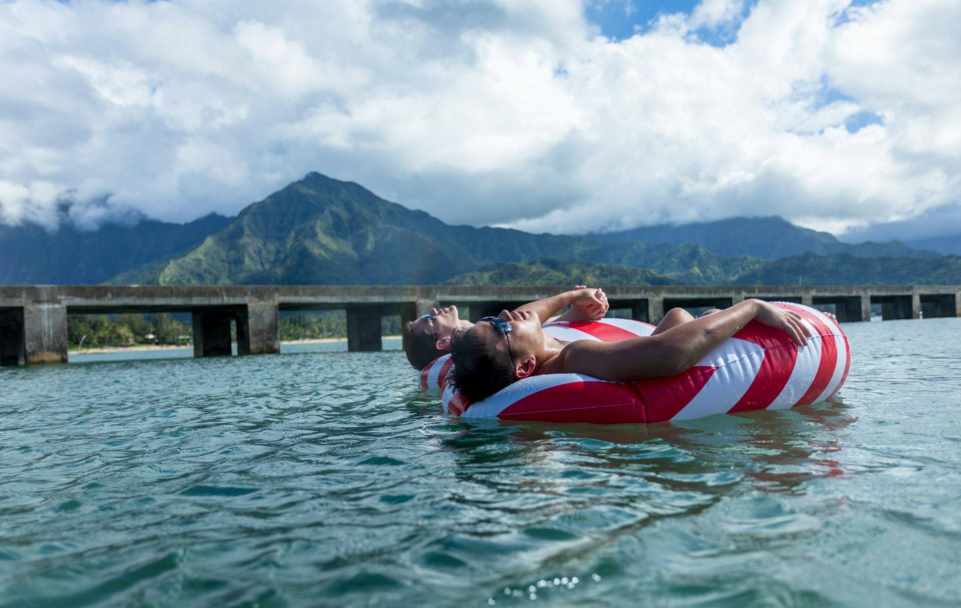 Two men holding hands and floating side by side in matching red-and-white-striped inner tubes on a cloudy day, with mountains behind them 