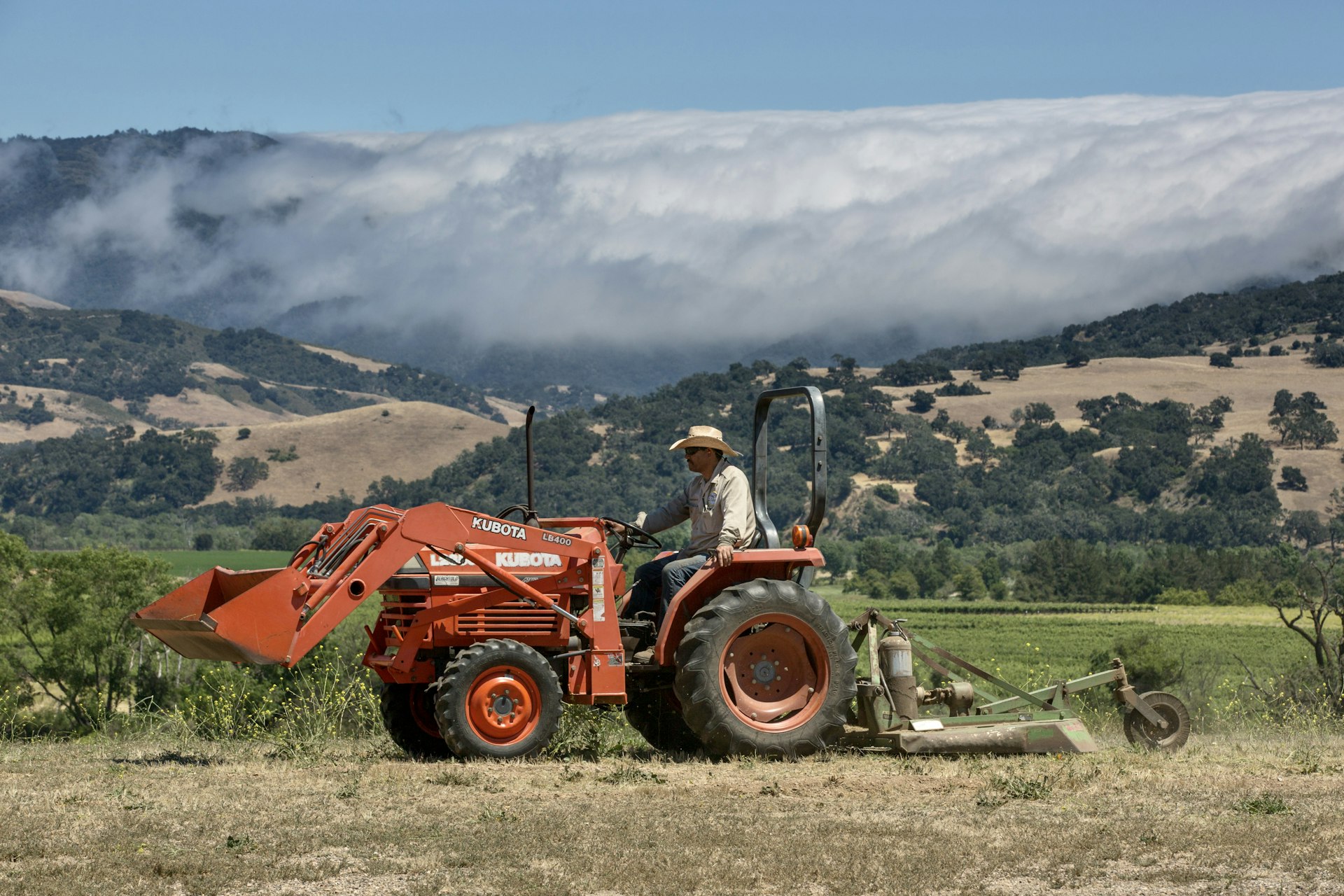 A farmer rides a tractor in fields, with a wall of fog atop of the Santa Ynez Mountain in the distance, Solvang, California, USA