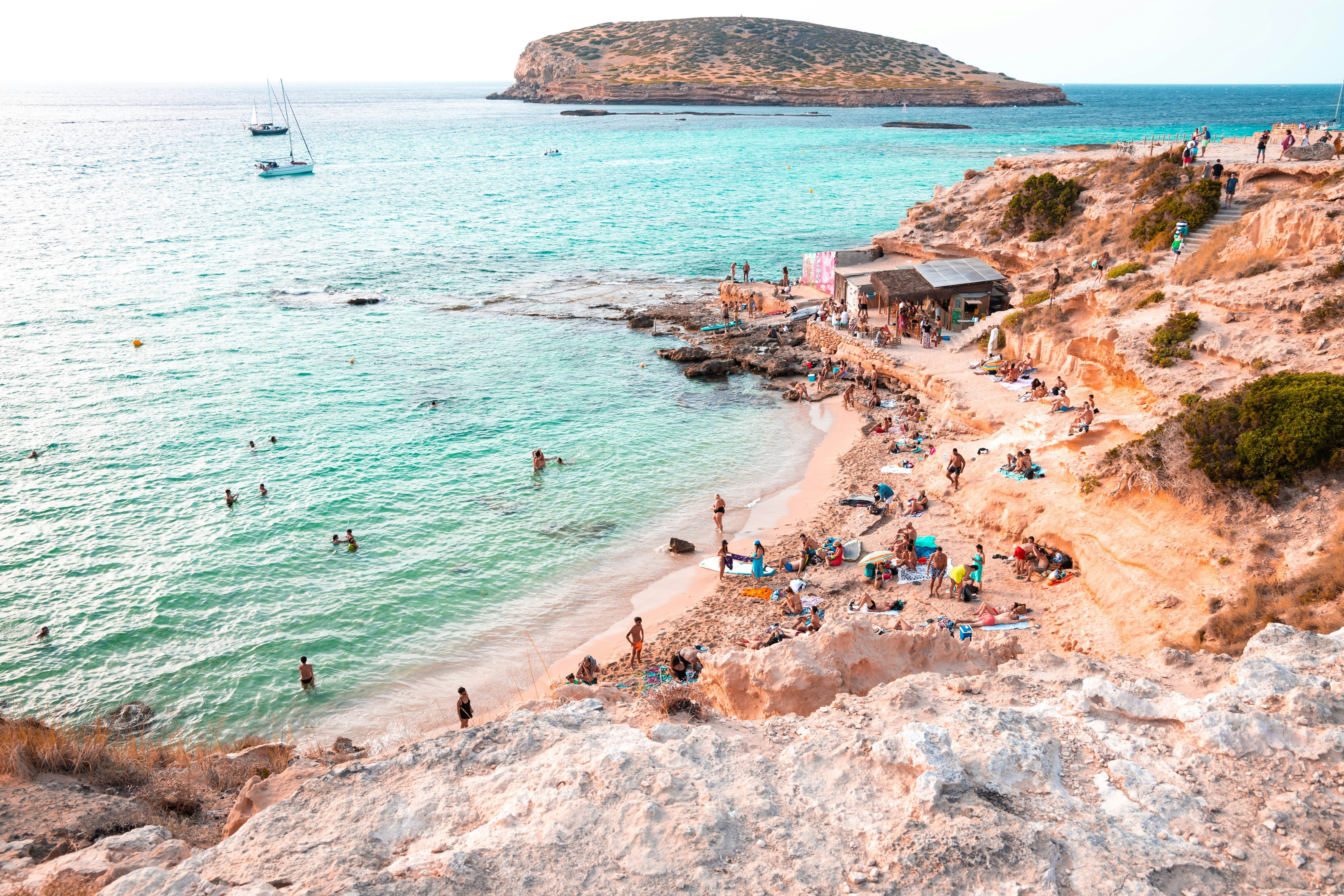 People line the sand at Cala Compte beach, Ibiza. A number of people sit on coloured towels while a couple swim in the sea.