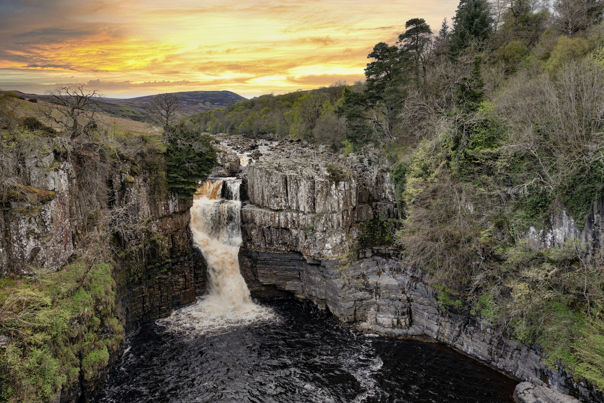 A waterfall backed by an orange sky cascades down a rock face into a pool