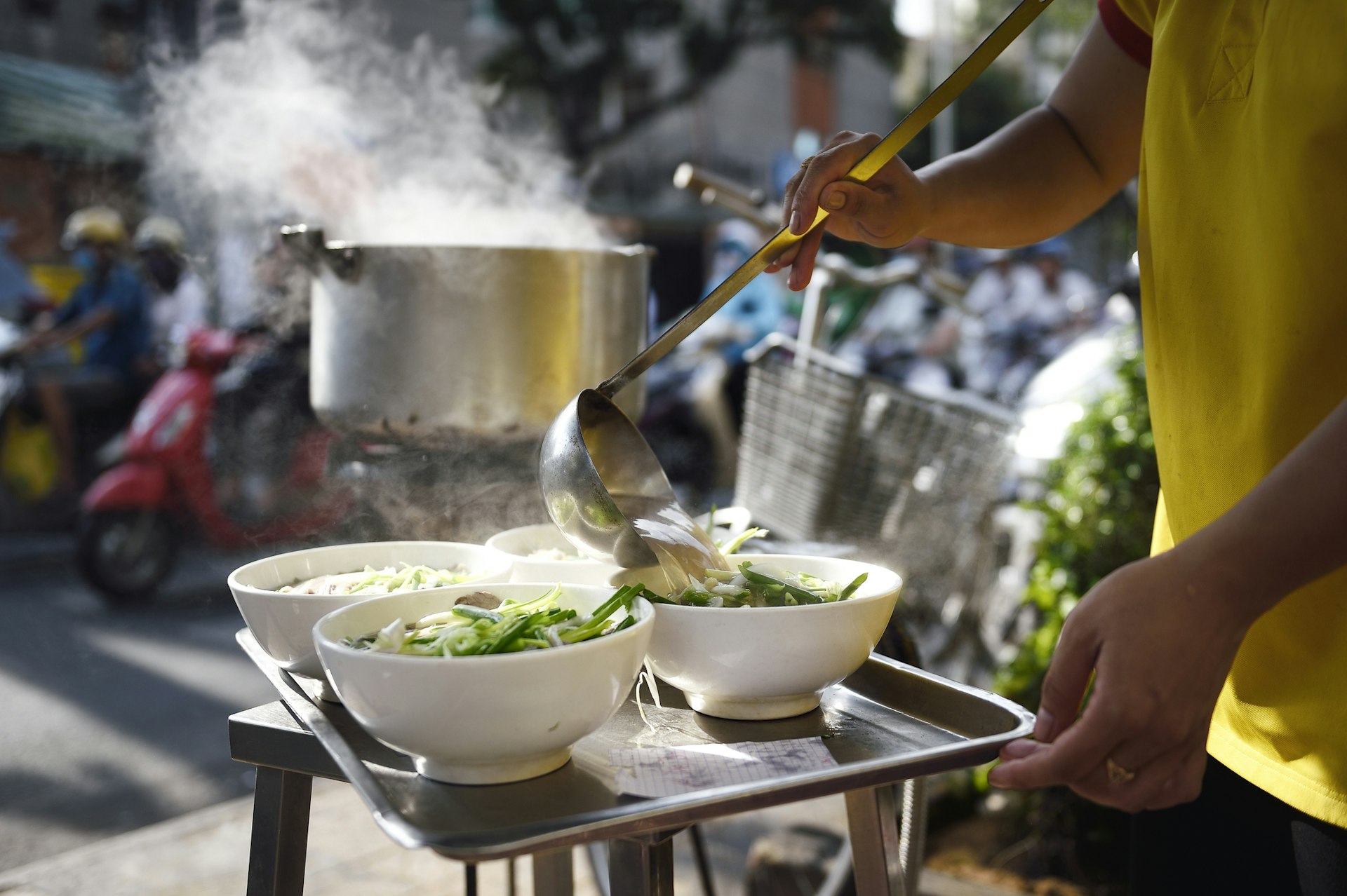 A hand ladels steaming soup into four bowls at a street food stall as motorcycles pass in the background