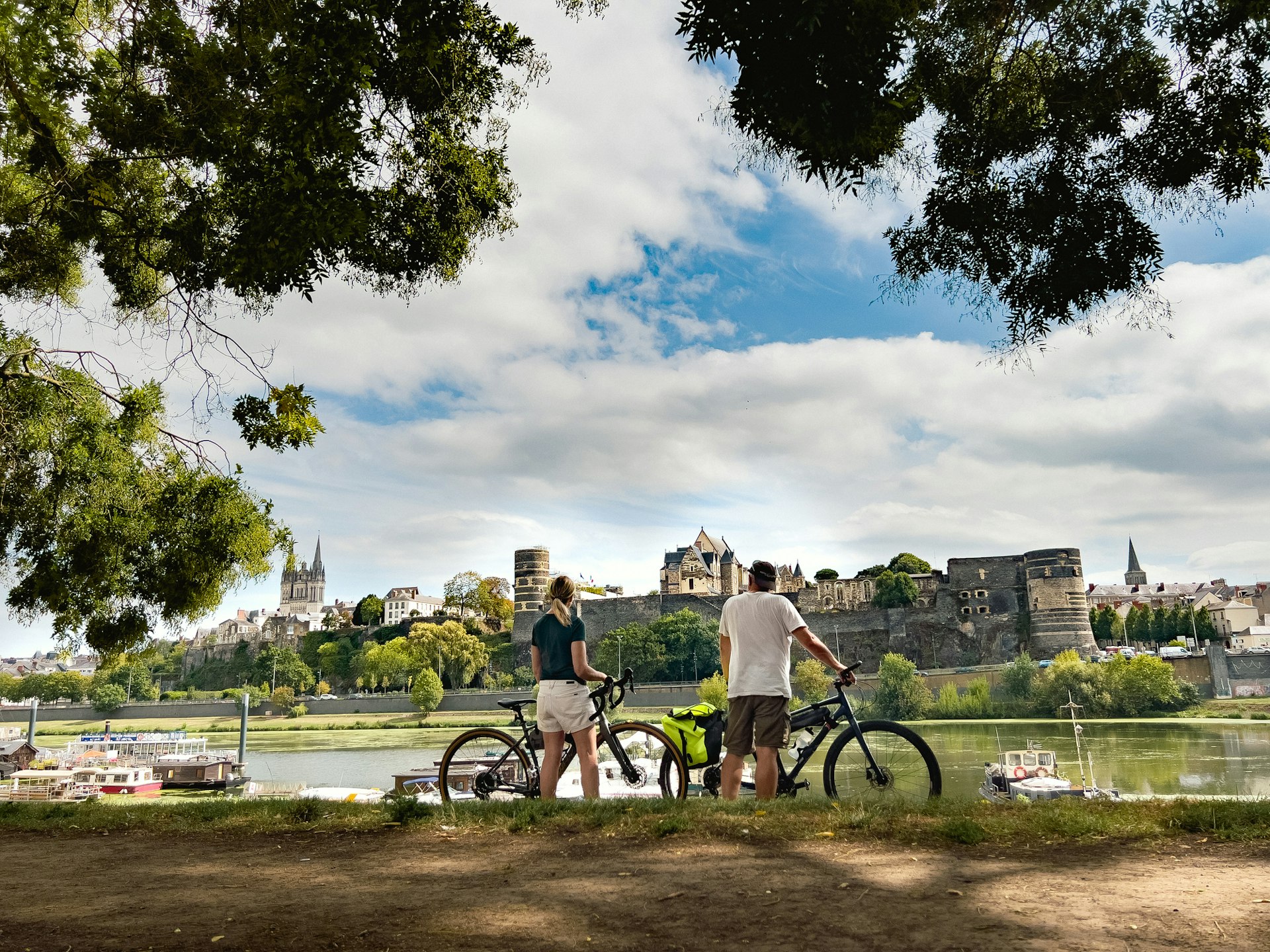 Two people pause riverside with their bikes to gaze across at a medieval city