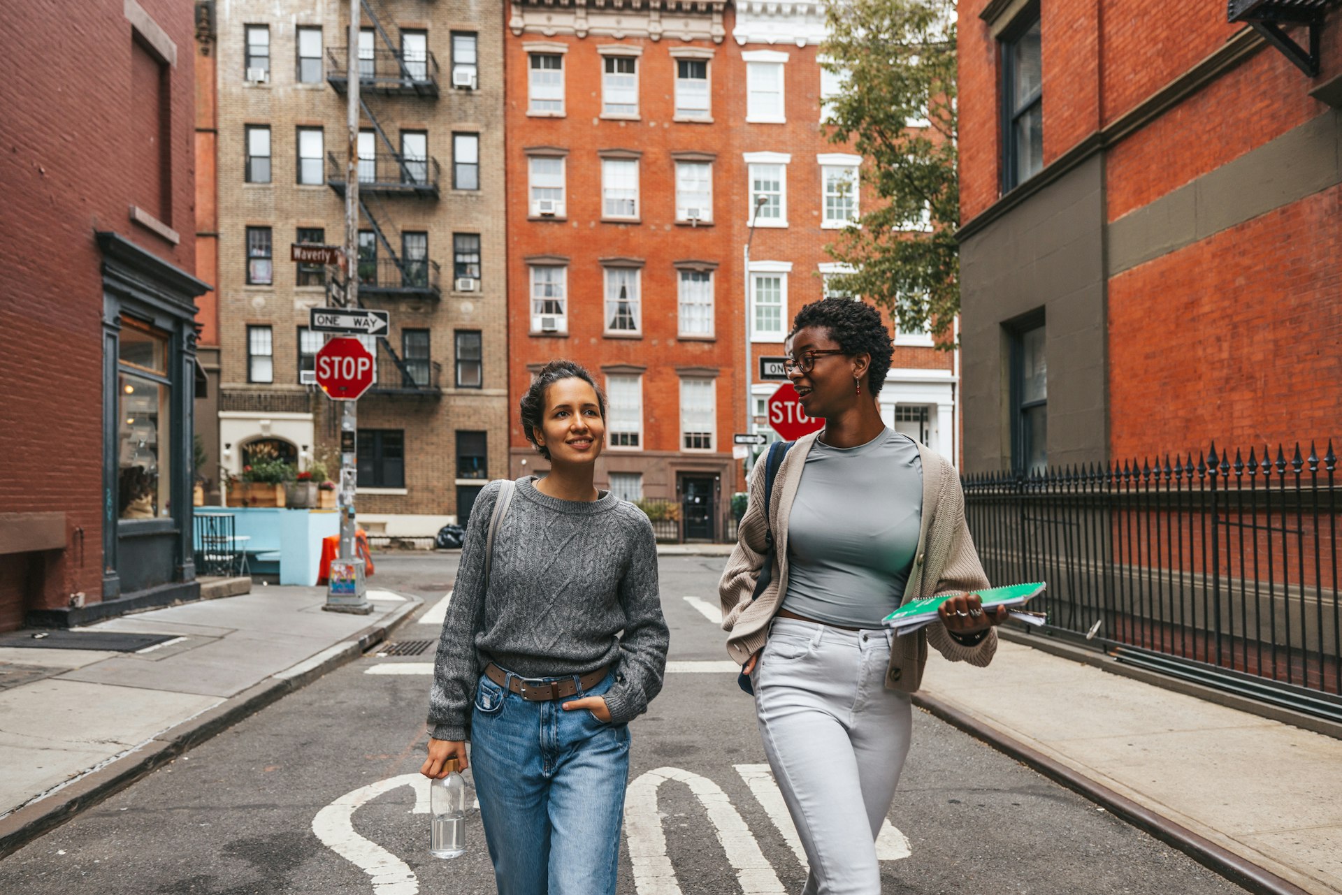 Two friends walk down a New York street with brownstone apartments in the background. 