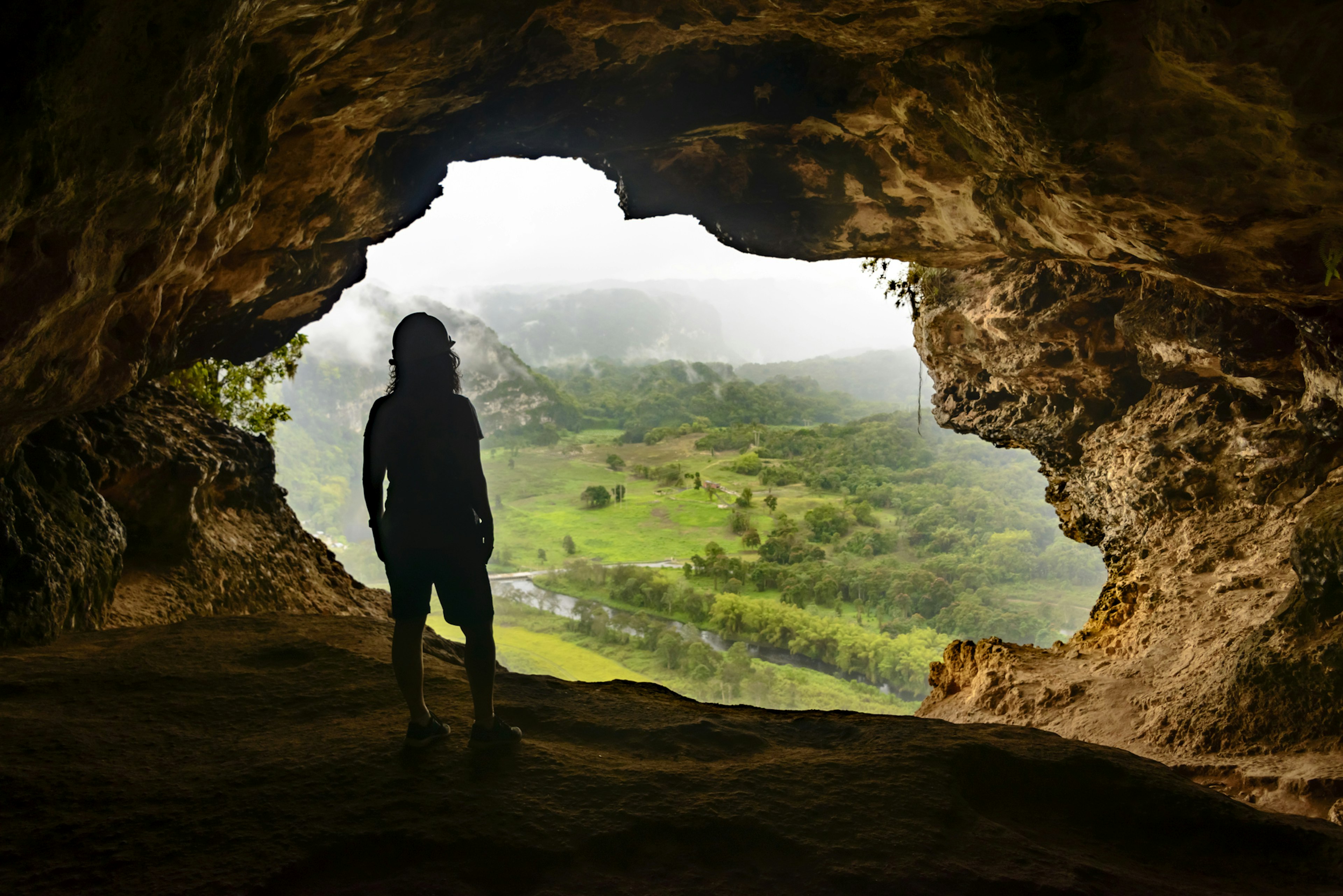 Person in silhouette looks out through the mouth of a cave to the green valley below