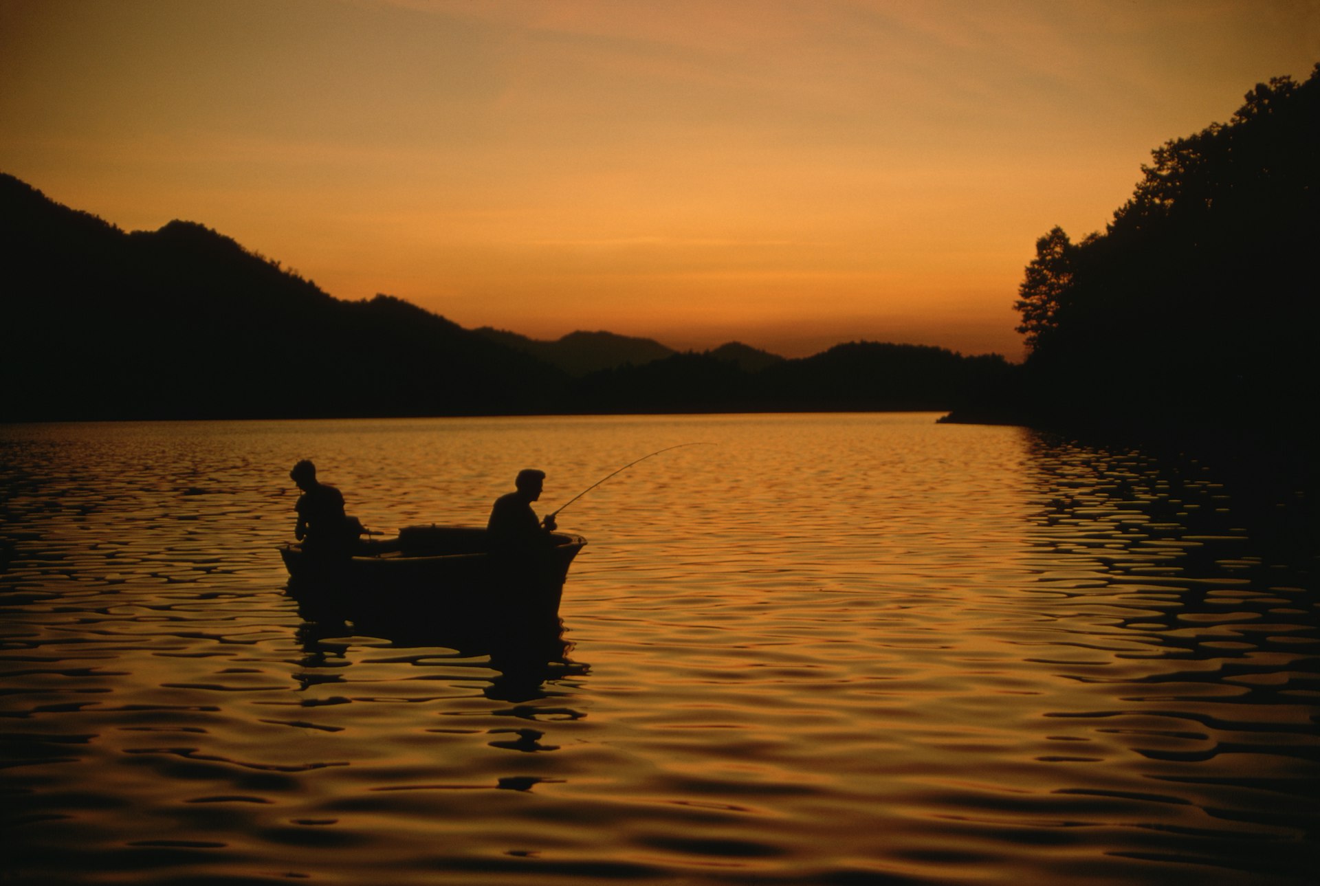 Two people in a small boat fish in a lake under an orange sunset