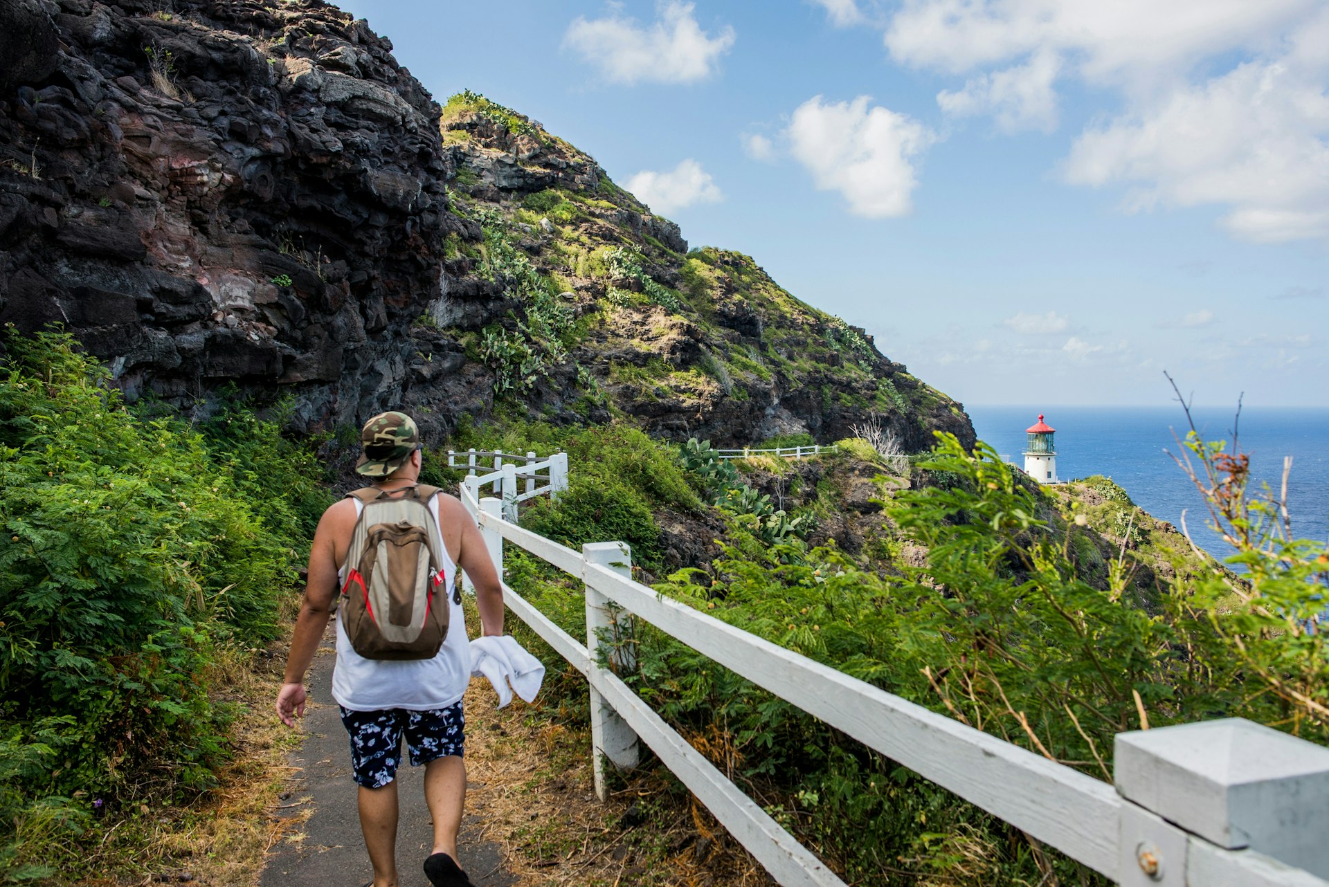 A white man in a baseball cap and shorts hikes along the Makapu'u Point Lighthouse Trail in Hawaii with the lighthouse itself in the far background, overlooking the sea