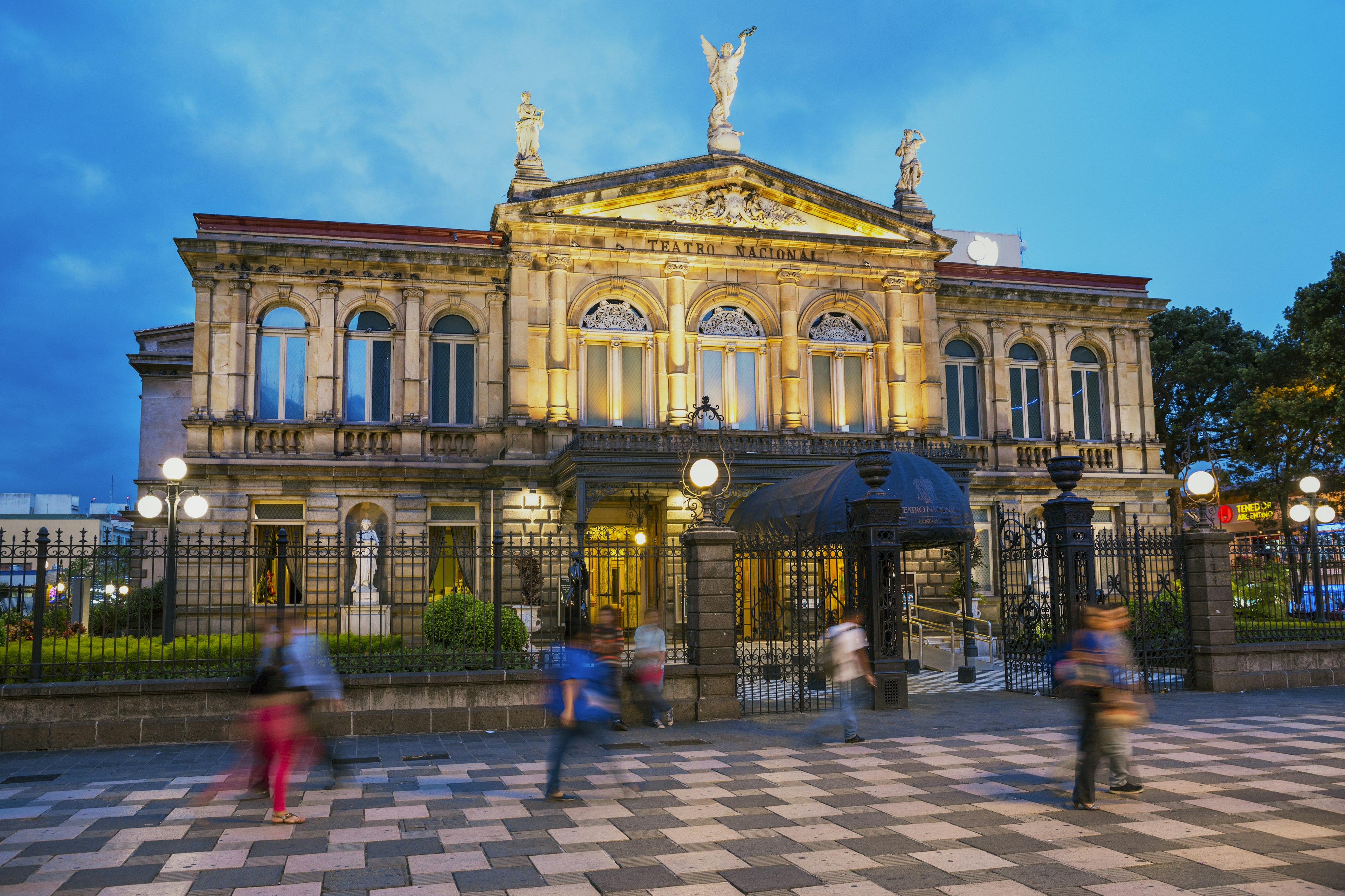 People walk in front of a grand theater building that's lit up at dusk