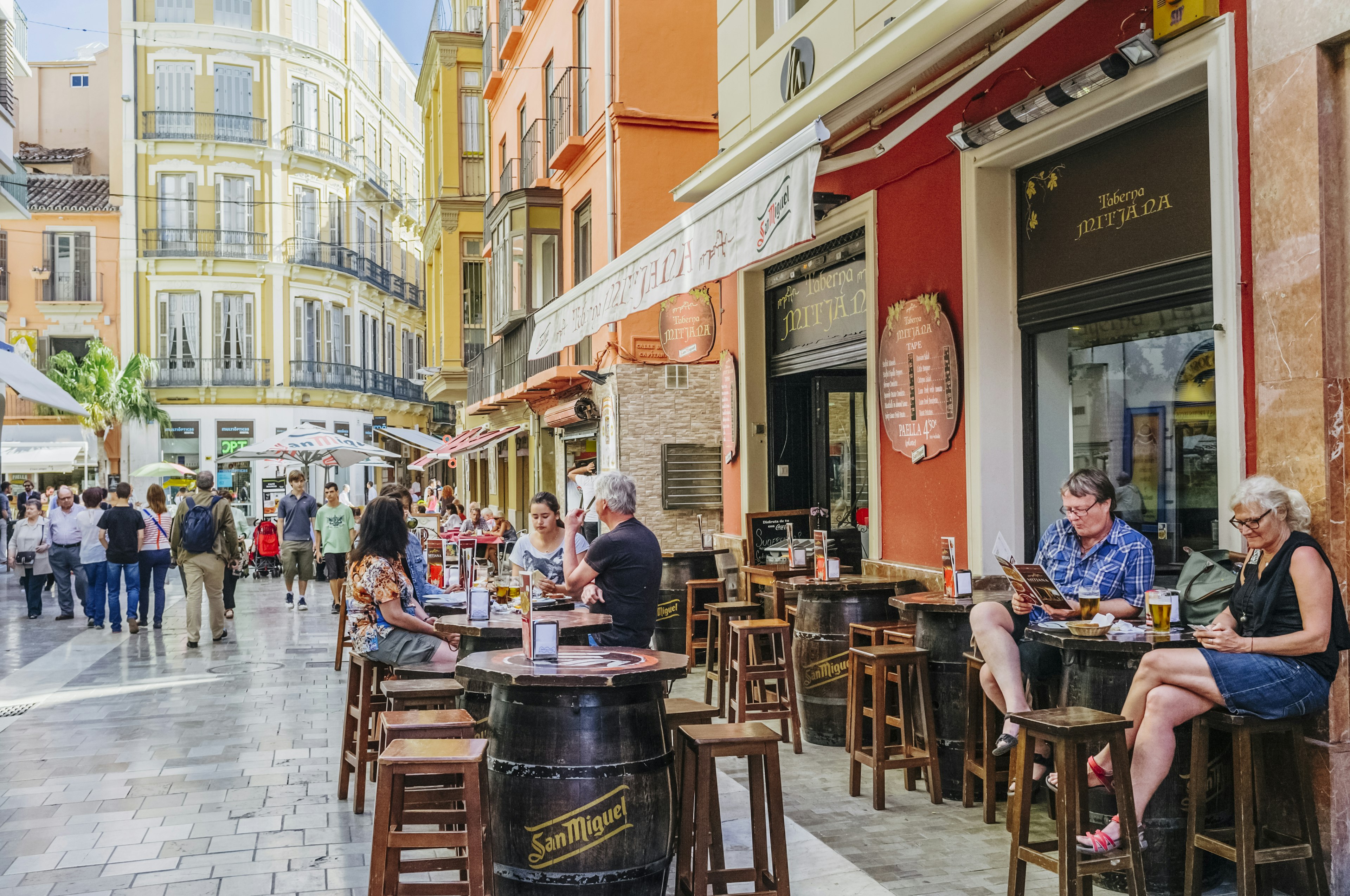 People sitting at the outdoor terrace of Taberna Mitjana bar in the historic center of Malaga, as tourists walk by.