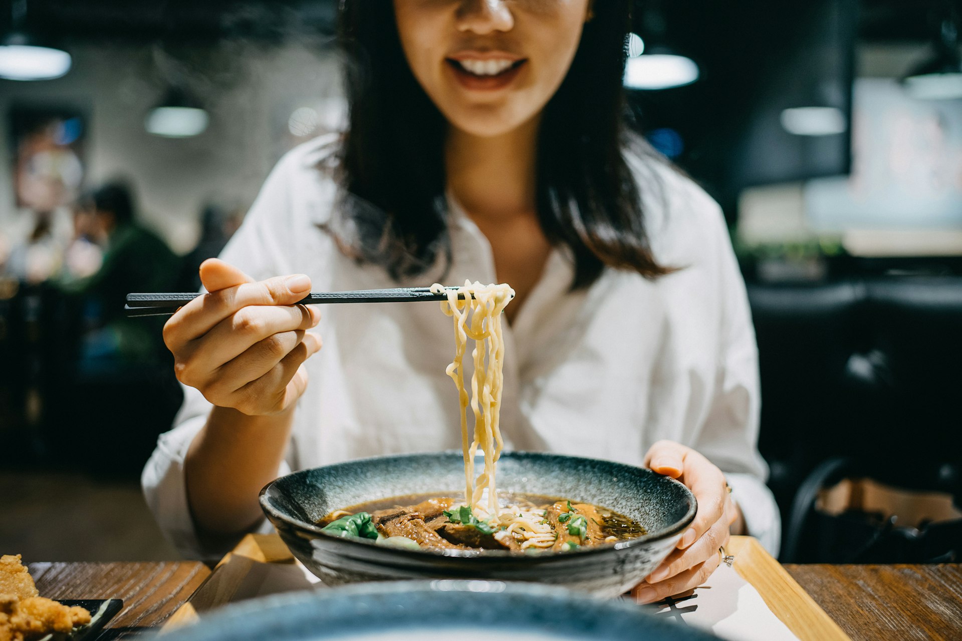Smiling young woman enjoying soup noodles with side dishes at a restaurant 