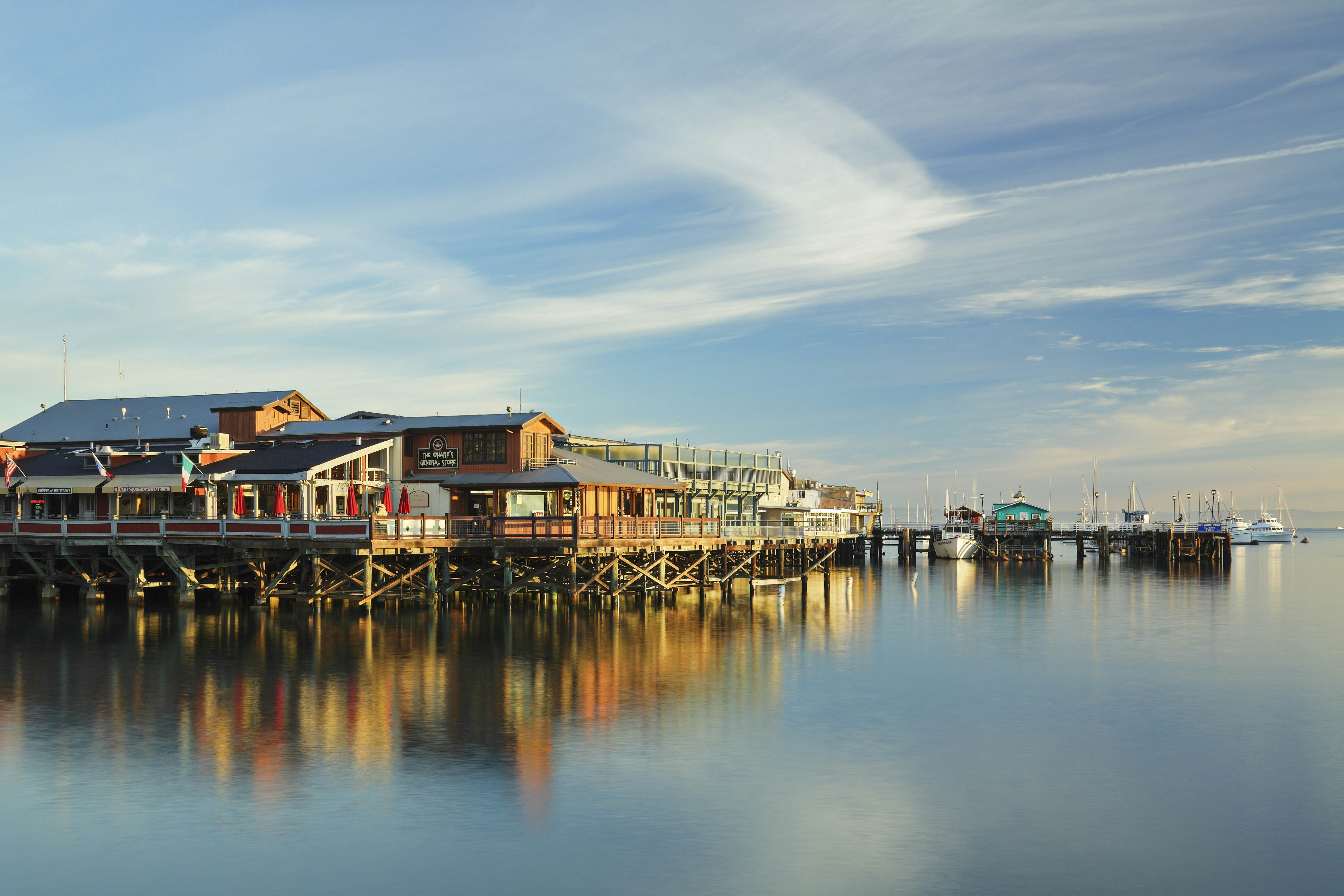 Wharf buildings reflected in the still water of the ocean and lit by an afternoon glow blue sky above dotted with clouds
