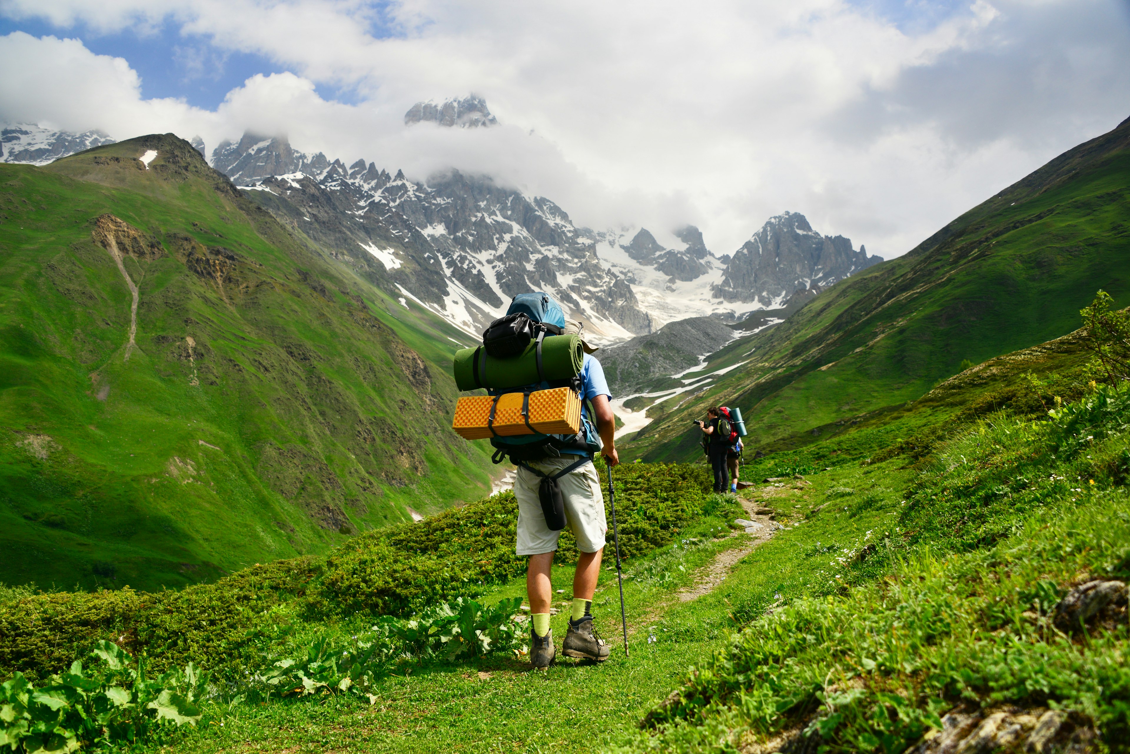 A hiker, laden with gear, follows a path in a mountain range. The hills are green with snow on the peaks.