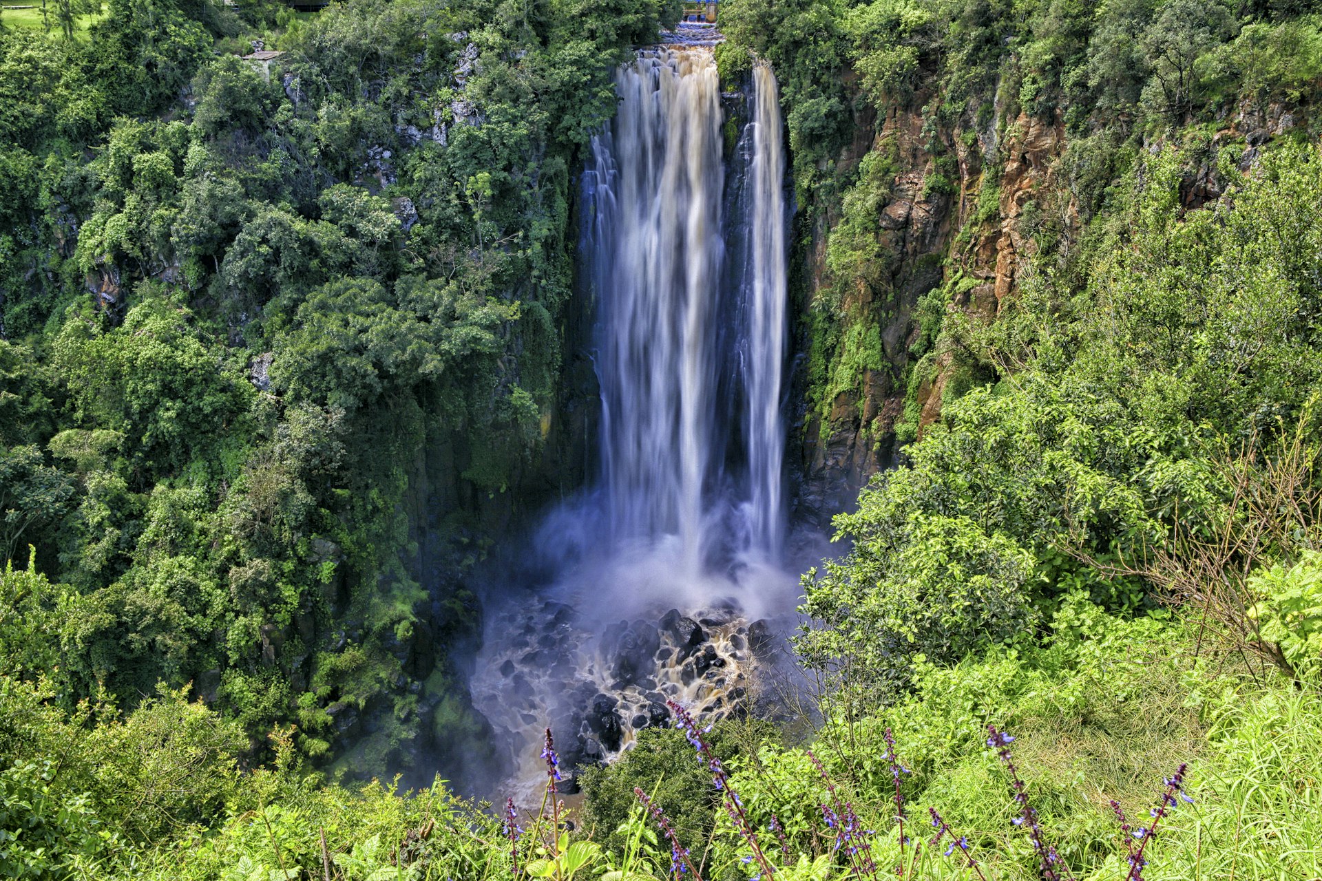 A shot of Thomson's Falls in Aberdare National Park from afar, the waterfall gushing down into a pool that's surrounded by thick greenery