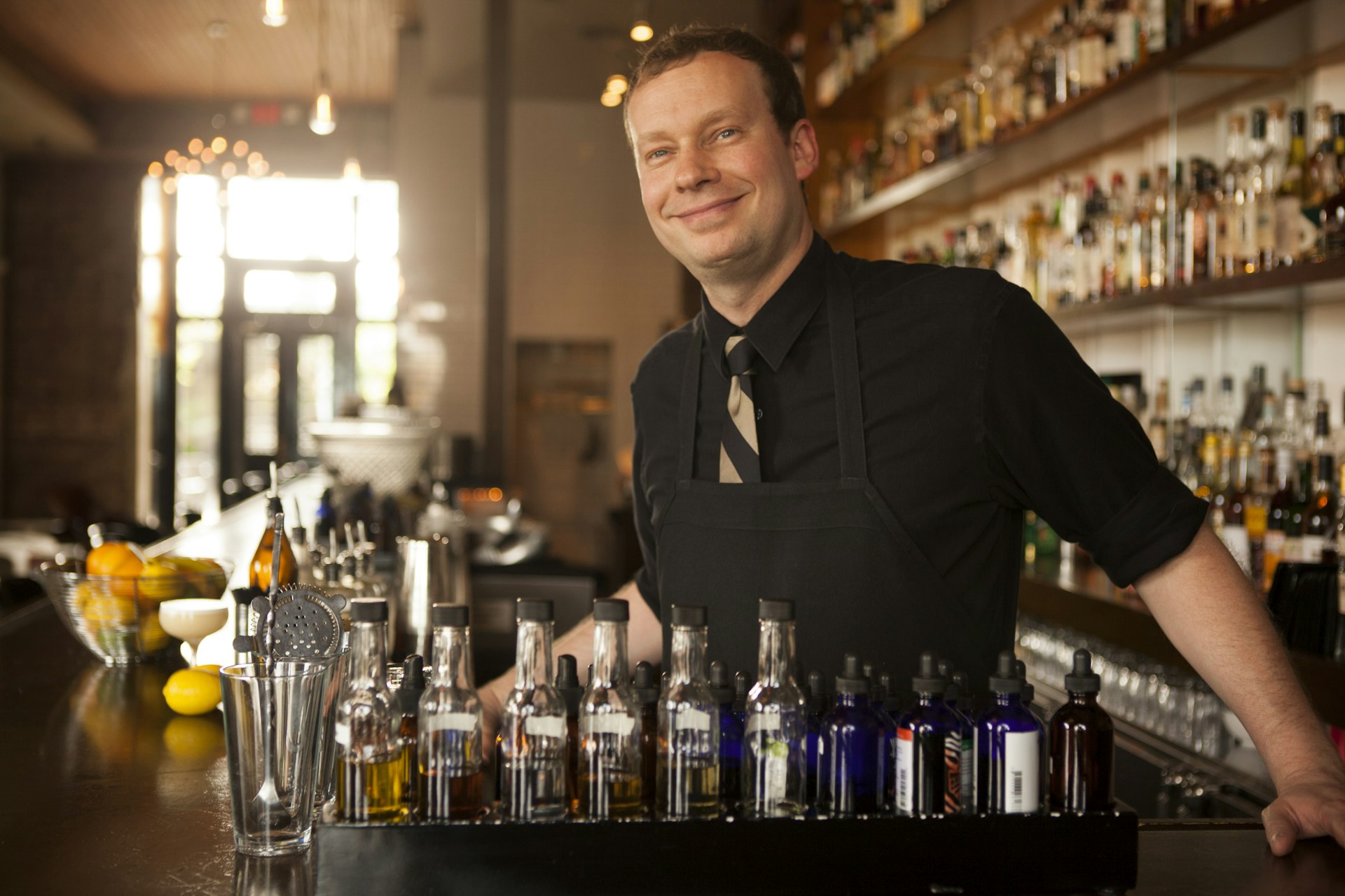 Portrait of a smiling barman at Cure bar in New Orleans with bottles lining the wood-panelled space