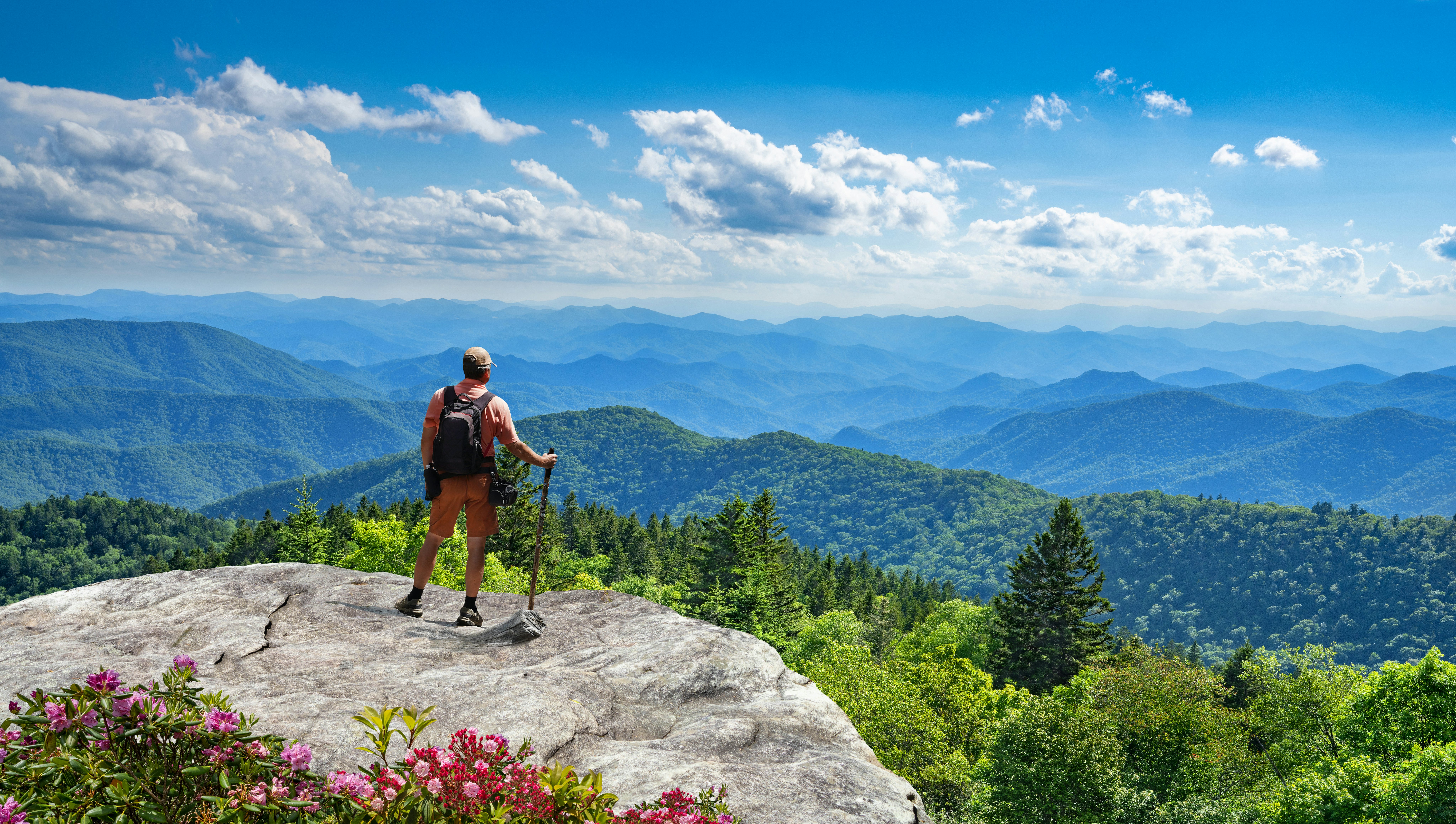 A man stands on a rocky outcrop on a mountain and looks out at a panoramic landscape of misty green hills and a blue haze in the distance.