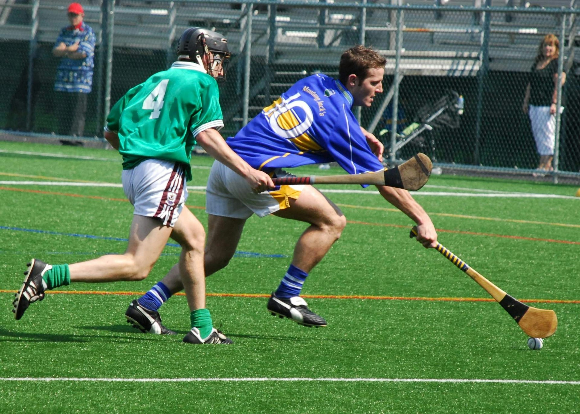 A hurling player in blue and gold gets to the sliotar in front of a green shirted player on a bright sunny day in Ireland