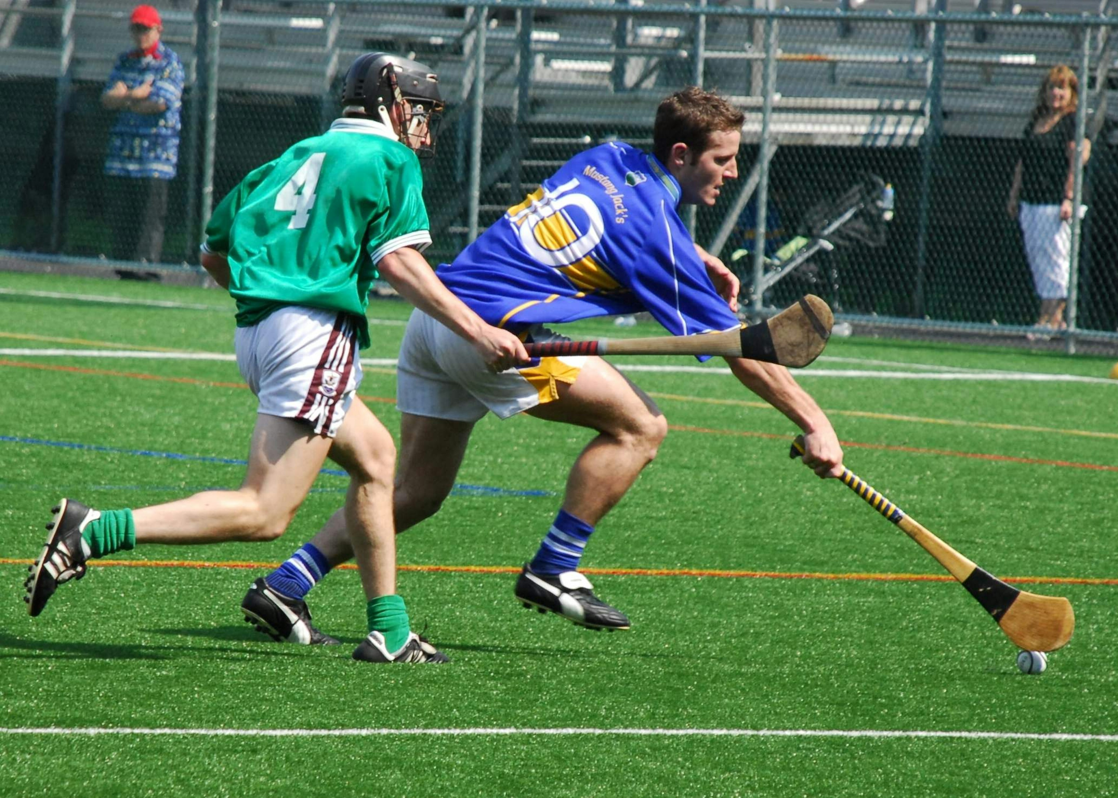 A hurling player in blue and gold gets to the sliotar in front of a green shirted player on a bright sunny day in Ireland