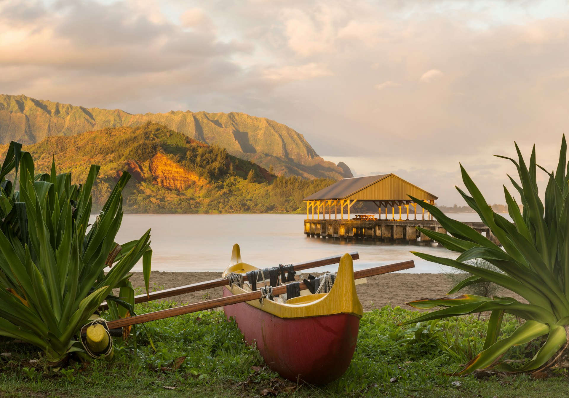 A wooden canoe sits on the edge of a beach with a covered pier, backed by mountains