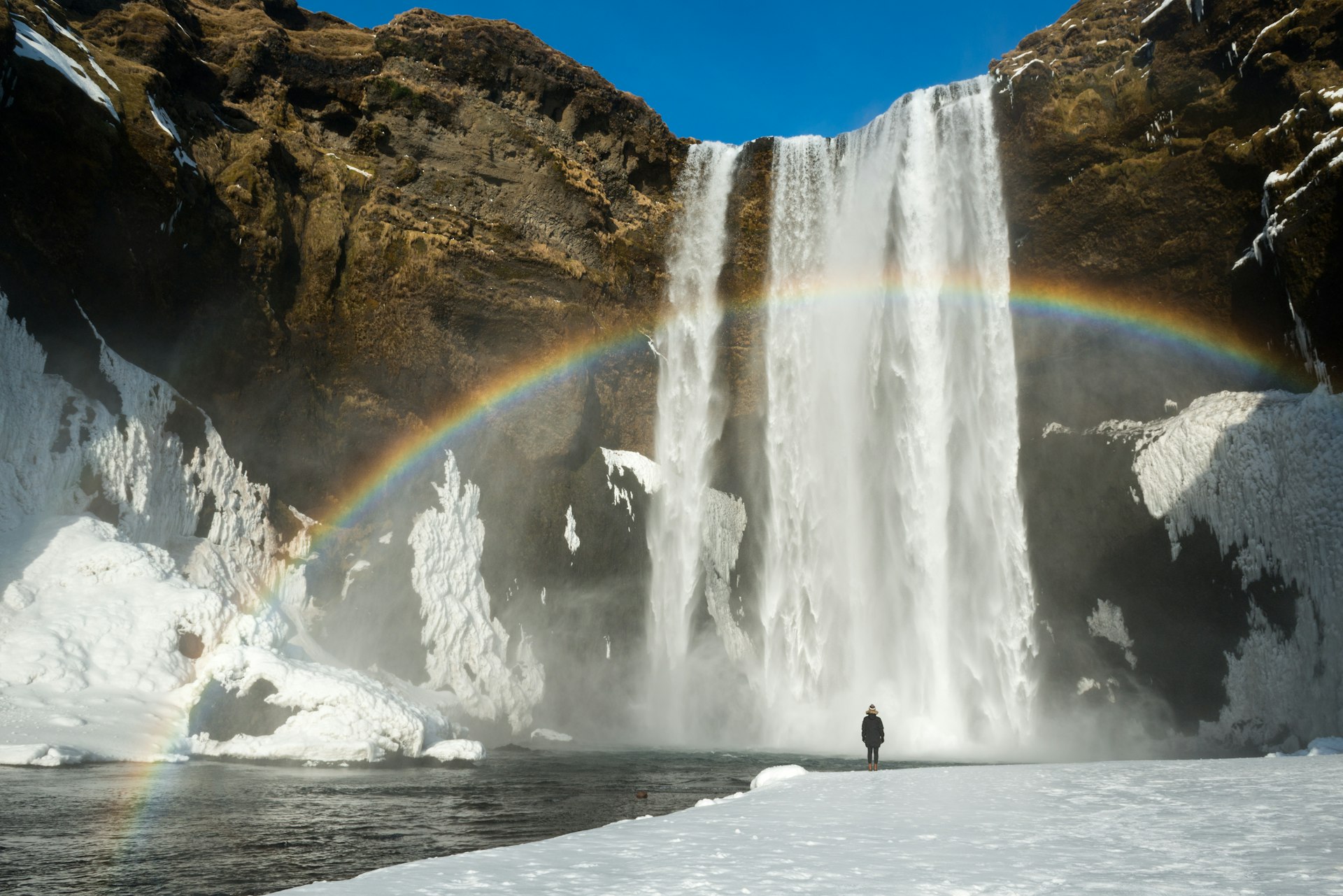 A tourist standing in front of Skogafoss waterfall with rainbow across it, Iceland