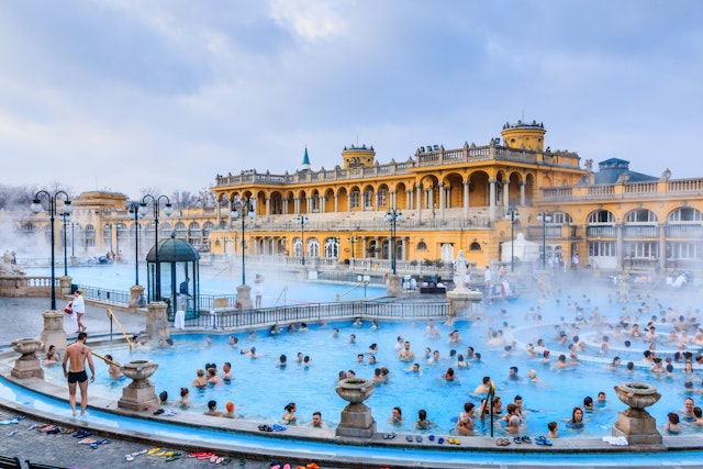 Bathers crowd in the bright blue Szechenyi Baths in Budapest with an ornate yellow building in the background.