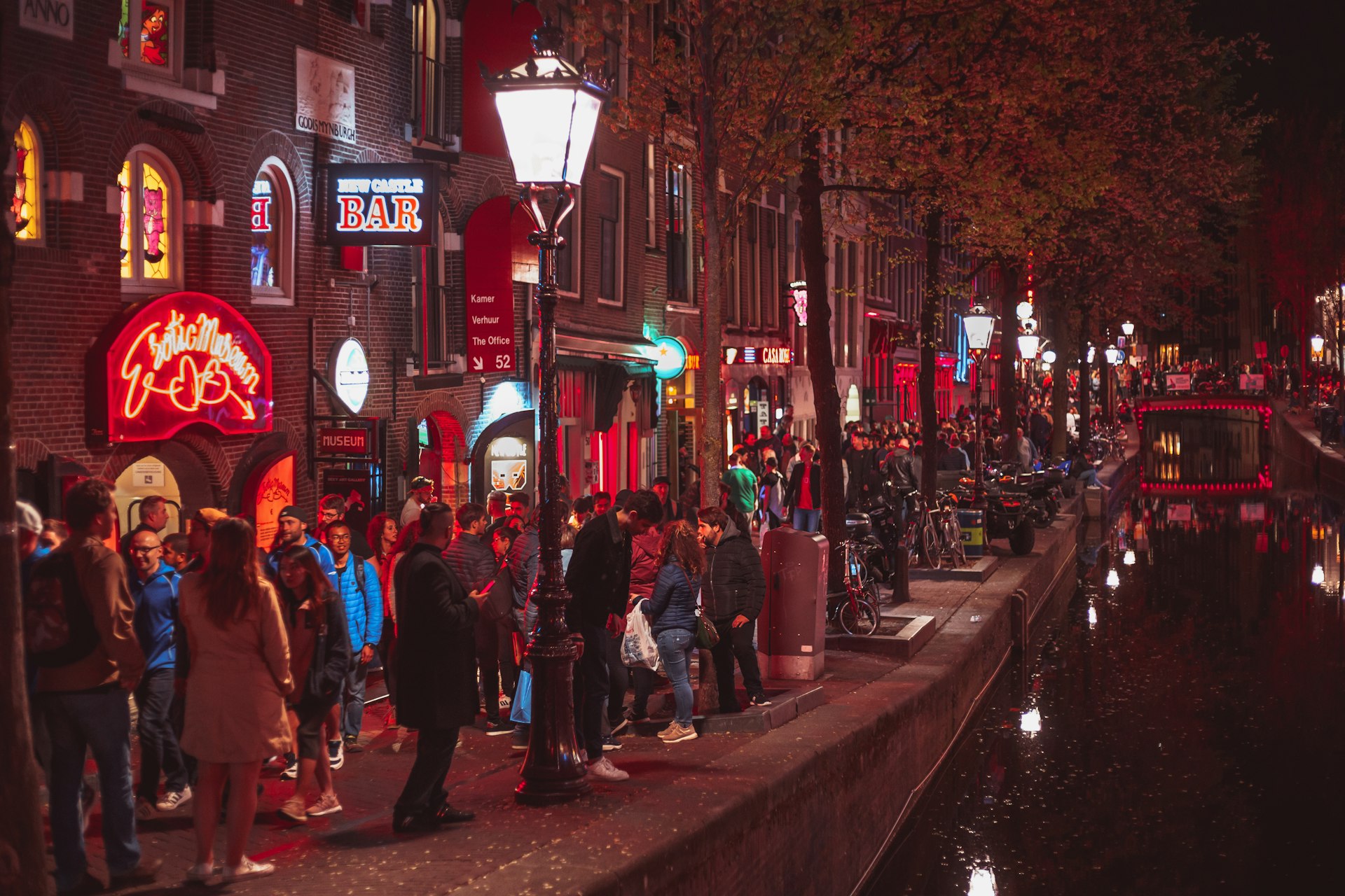 Crowds of tourists walk along a pathway between a canal and a row of bars and places advertising sex work in Amsterdam