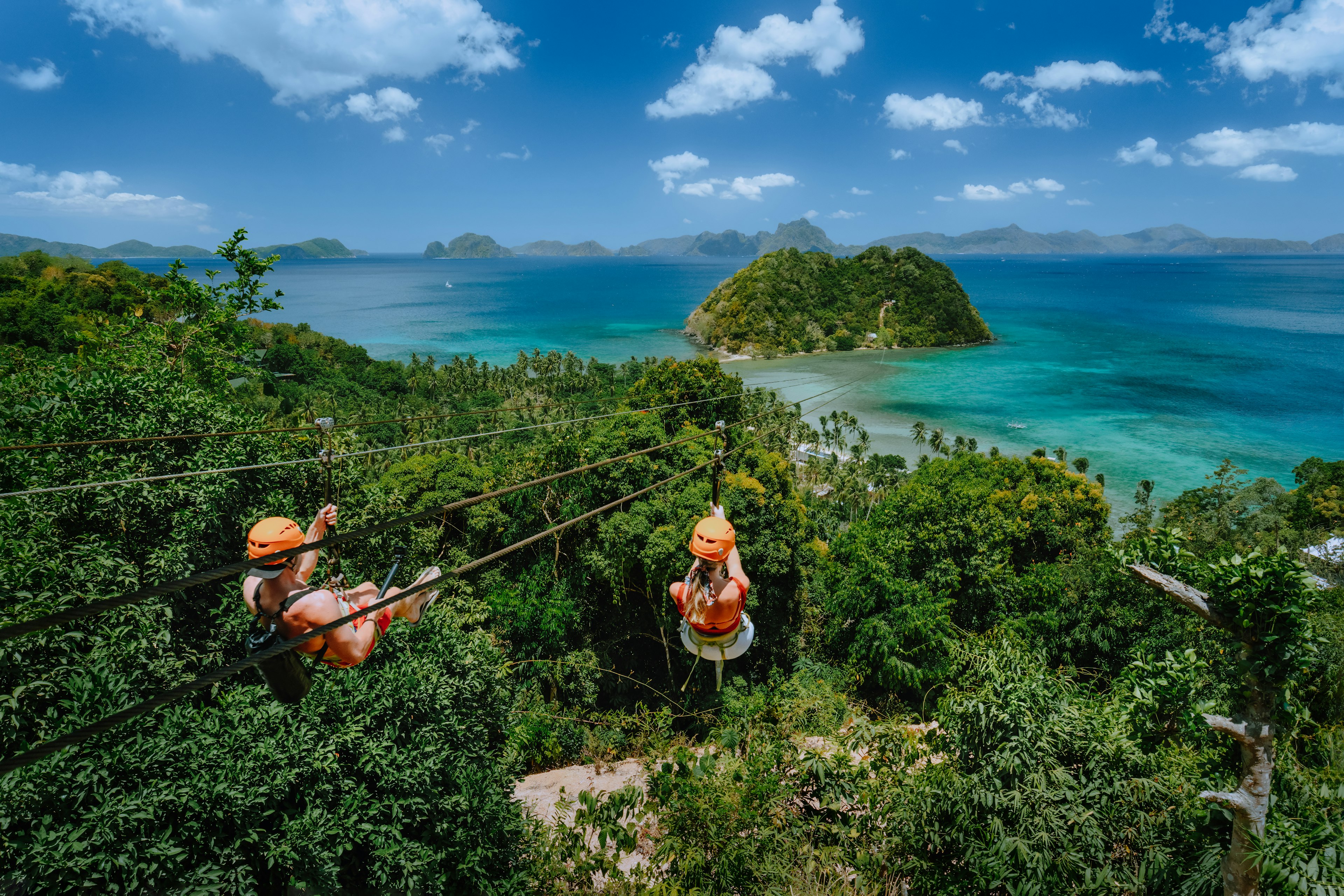 Two people in helmets ride a zip-line over a canopy of tropical trees toward the shoreline with bright blue water