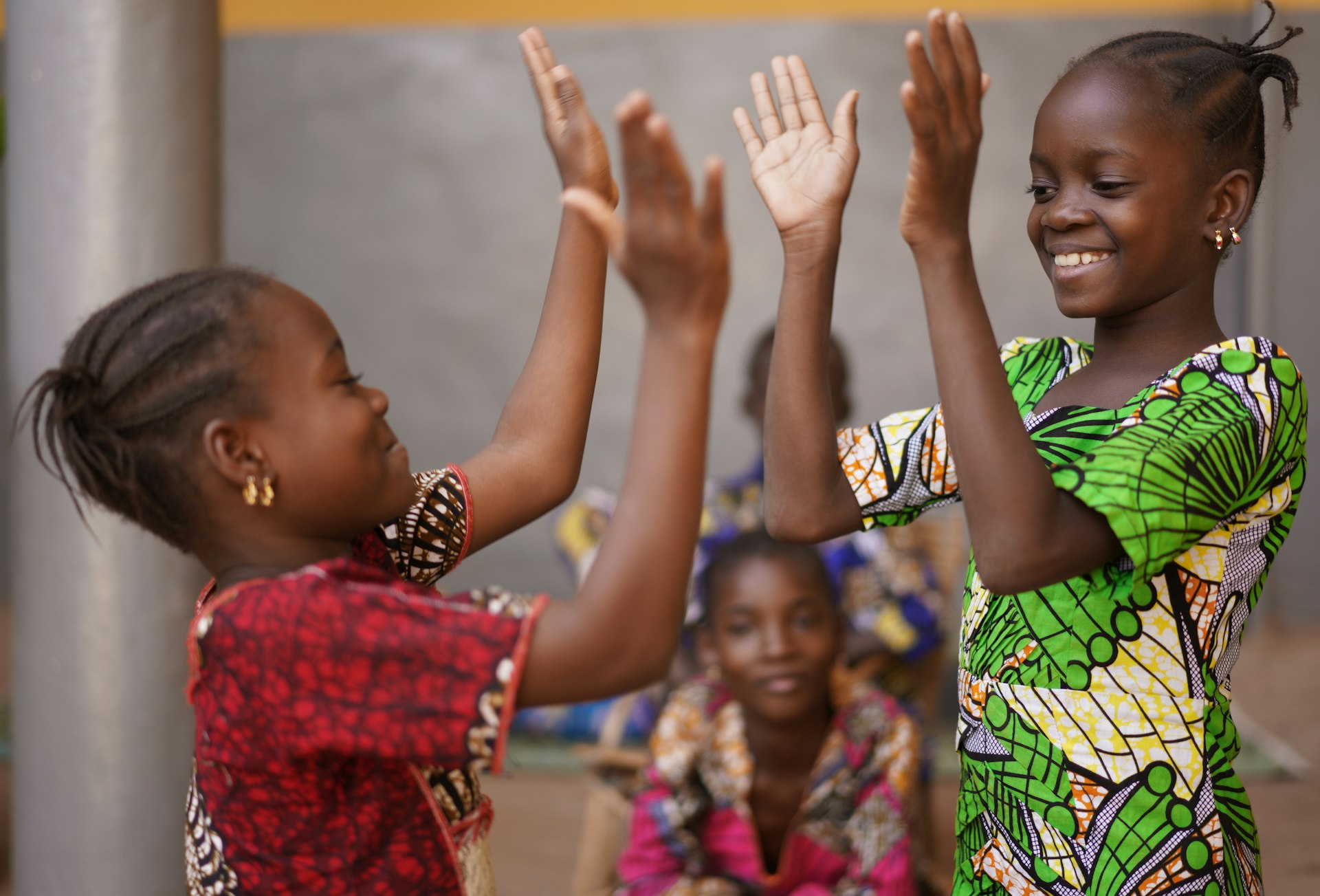 Two children in brightly colored traditional Nigerian dress smile as they play a hand-clapping game at a celebration