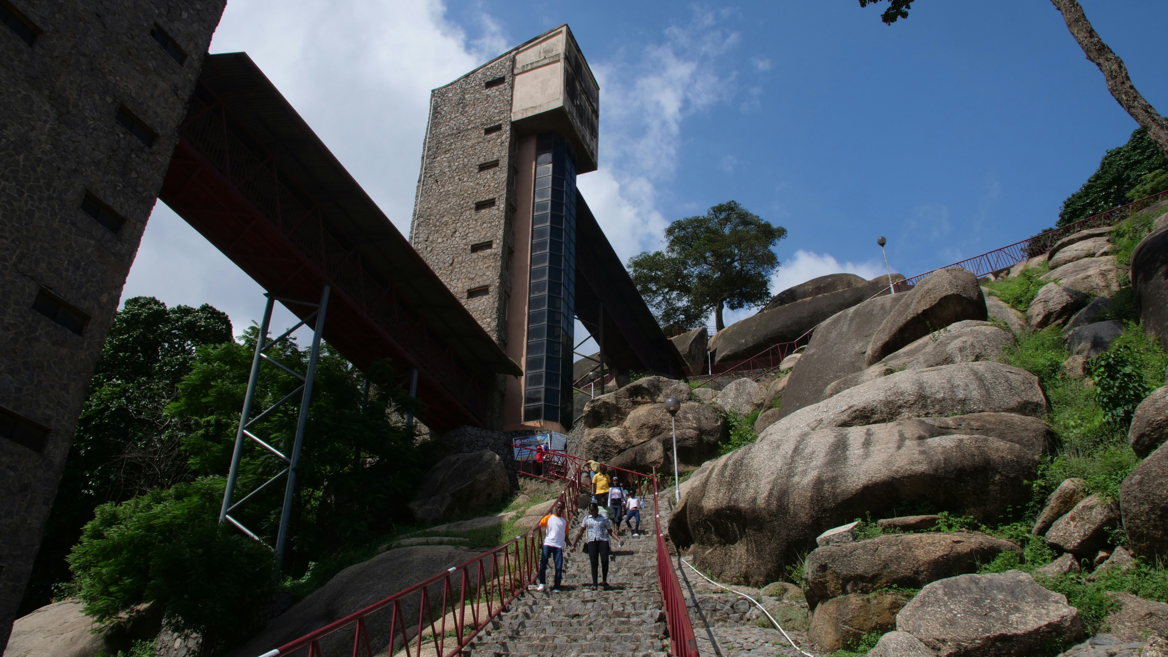 People hike up steep steps leading to a large rock formation on a hill