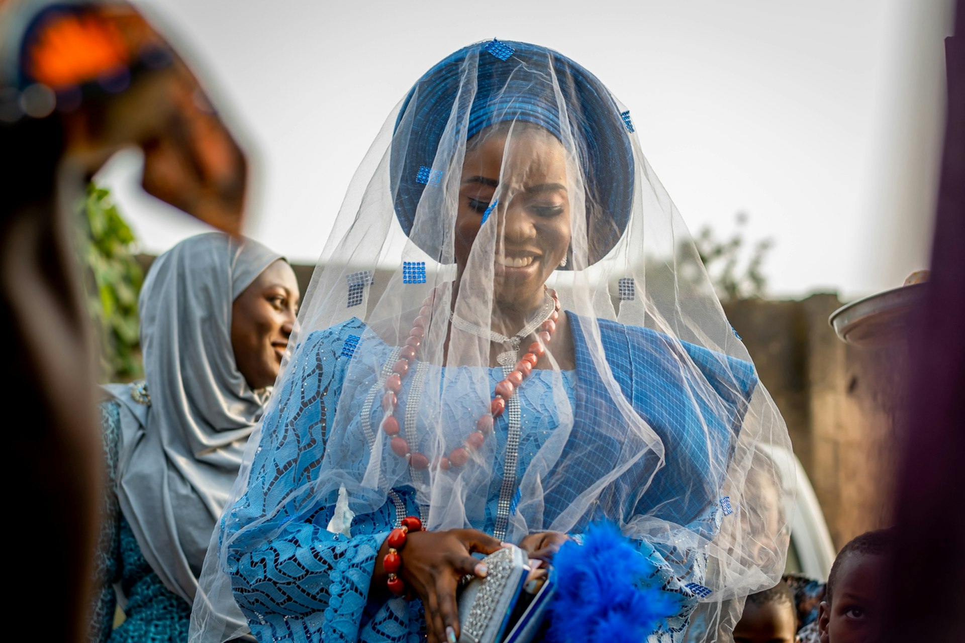 A woman dressed in blue with a light veil covering her head and face smiles at a wedding celebration