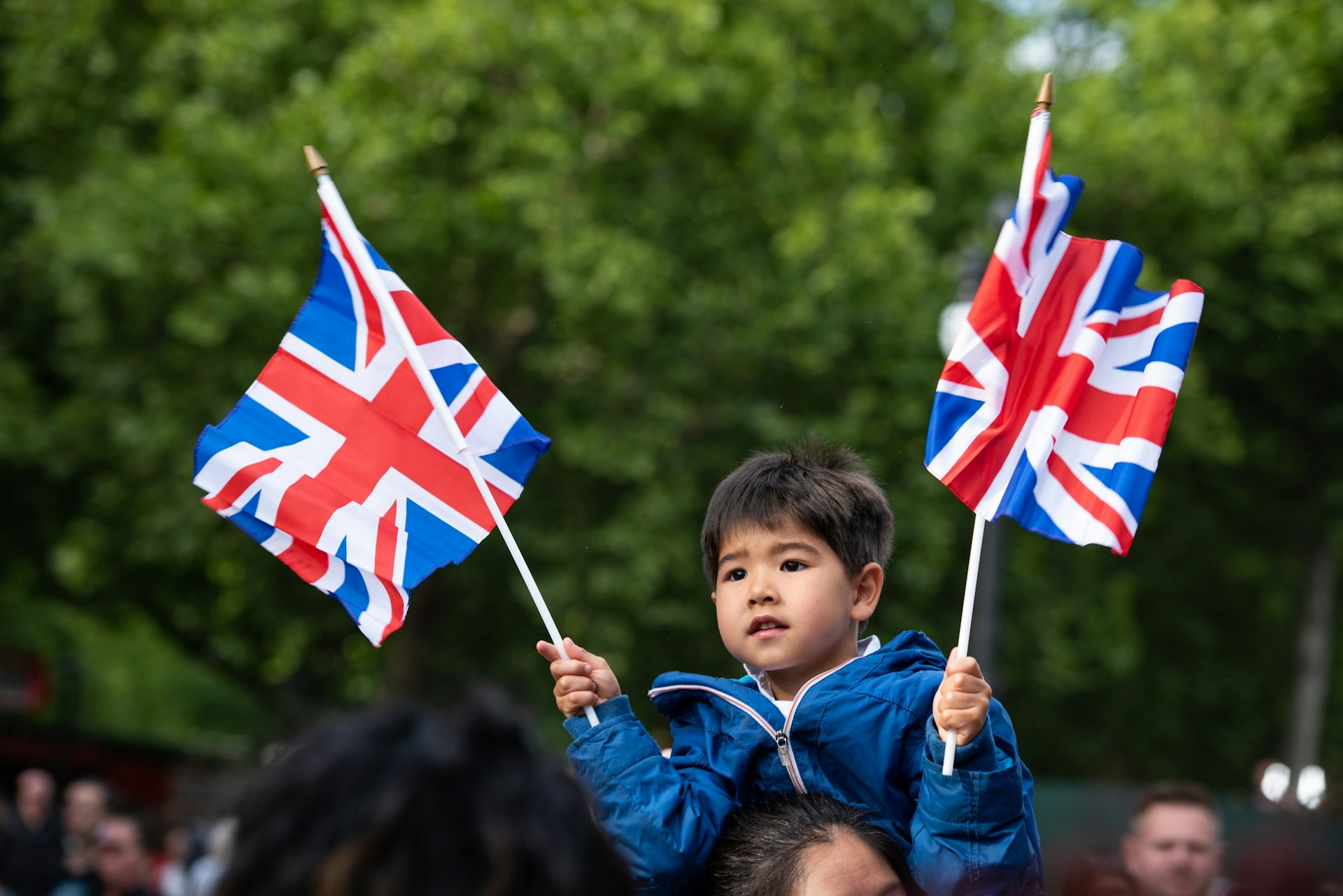 A boy sits on his father’s shoulders waving two Union Jack flags in London, England, United Kingdom
