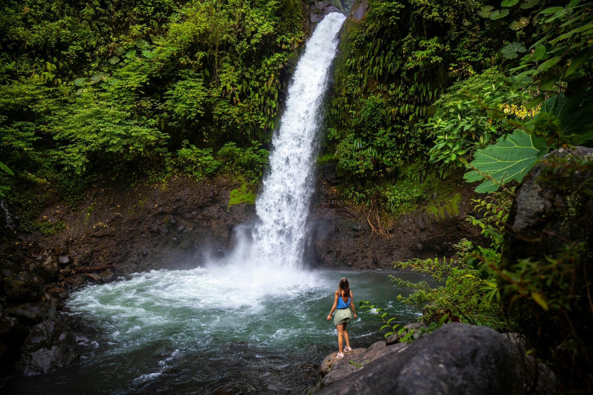 A woman stands on a rock at the edge of a pool with a waterfall plunging into it