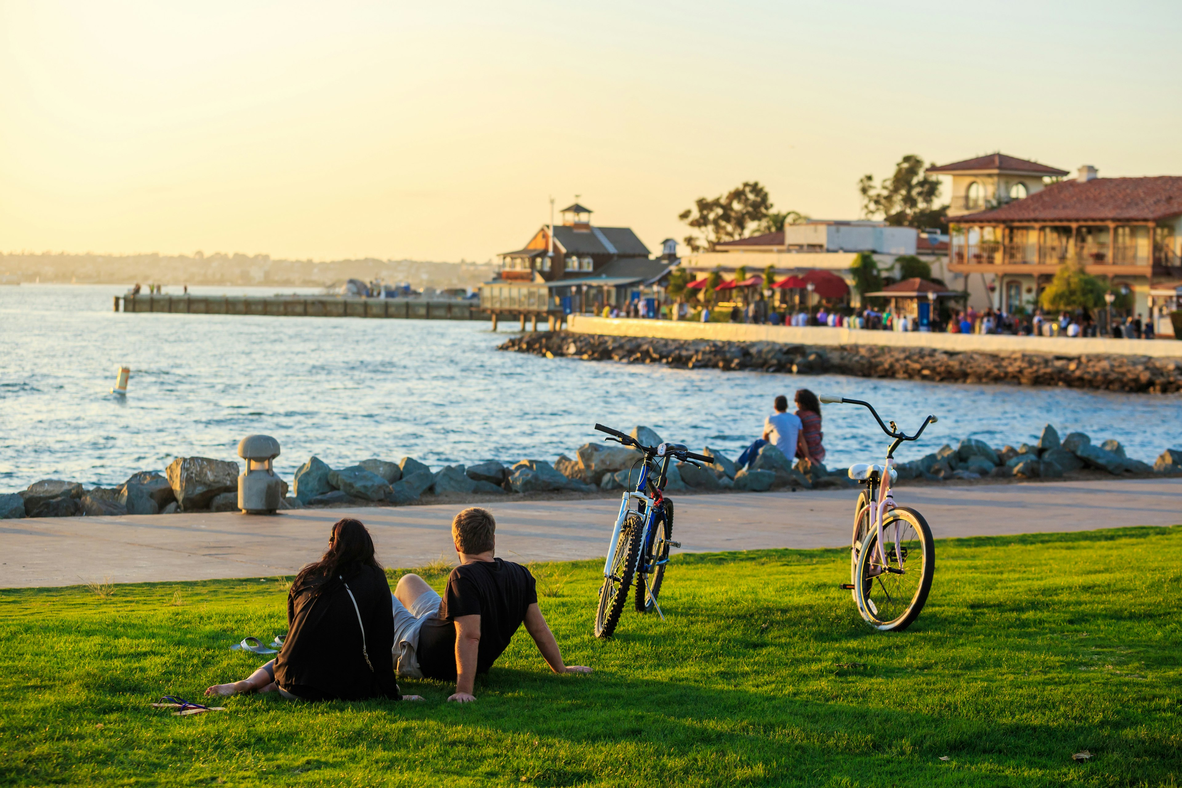 People sit on the grass near a waterfront as the sun is setting