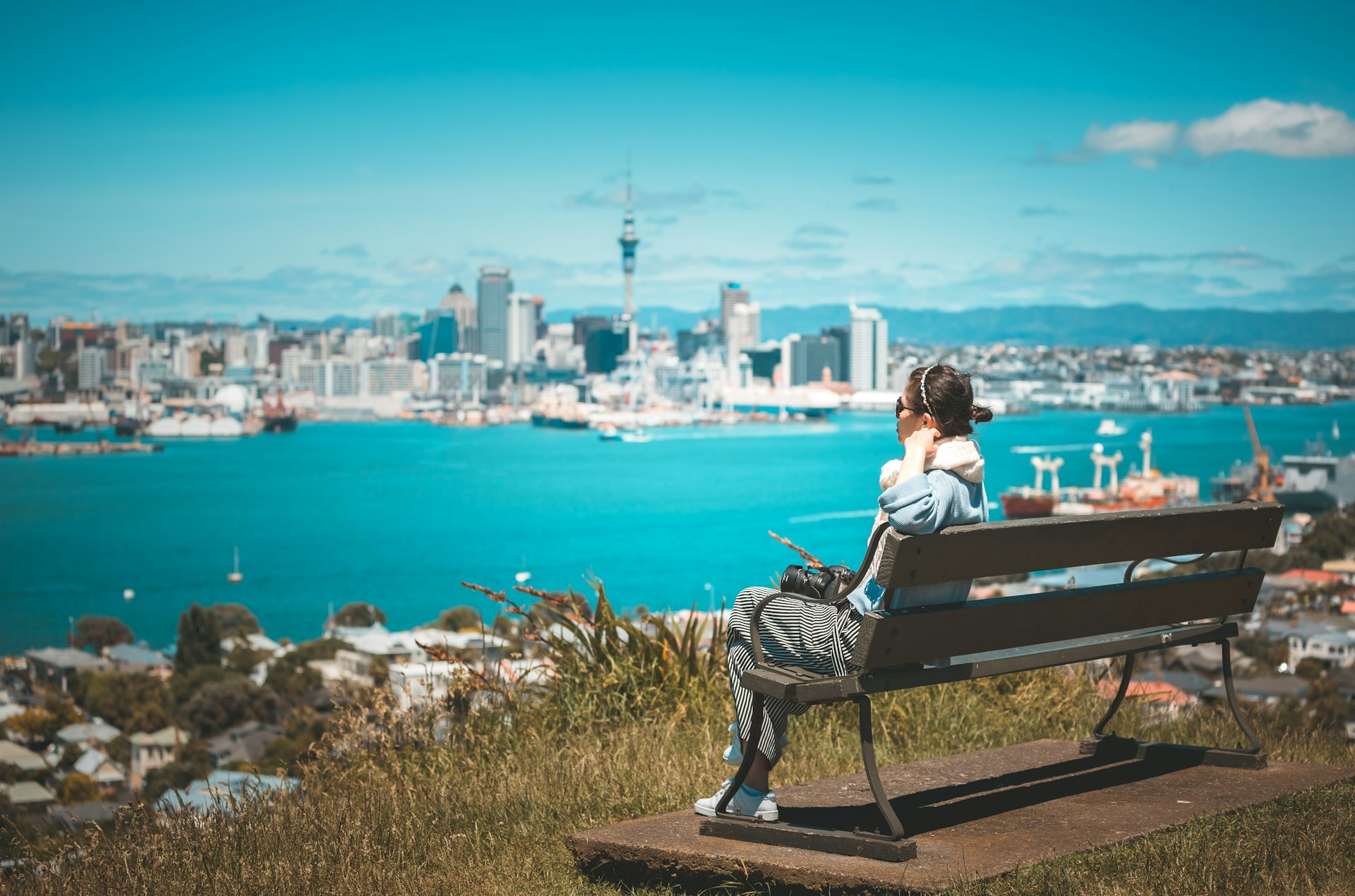 A woman sits on a bench at a viewpoint looking across a body of water towards a city skyline