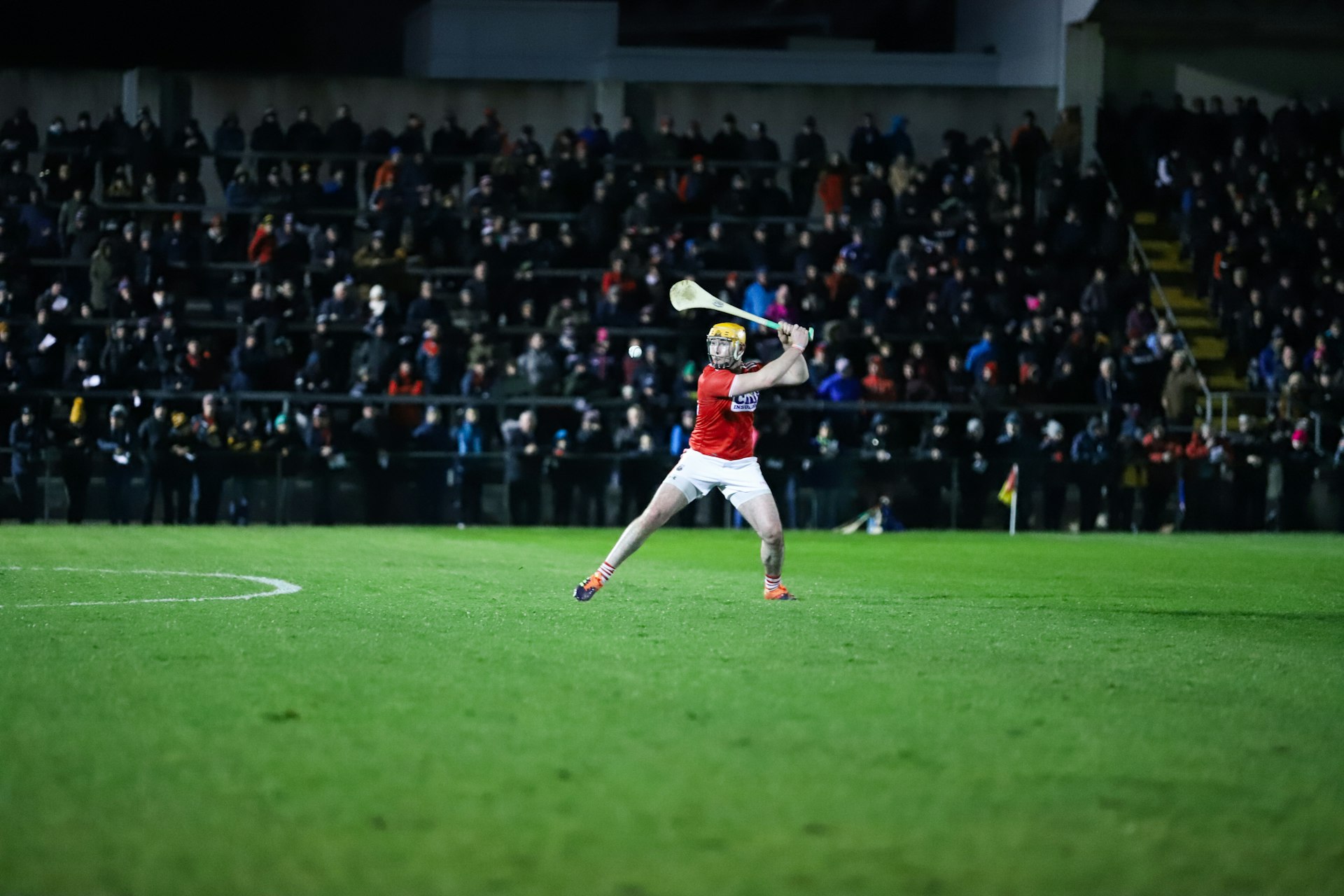 A hurling player in red prepares to hit the sliotar in front of the blurred crowd at Mallow GAA Sports Complex in Irleand