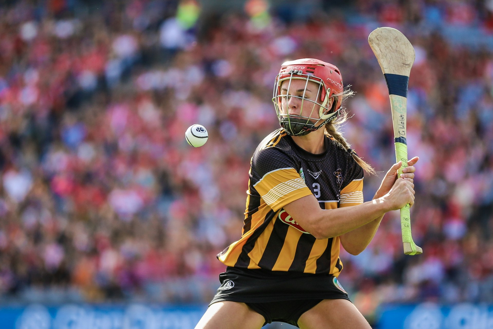 A helmeted woman Camogie player in gold and black stripes prepares to hit the sliotar during a game