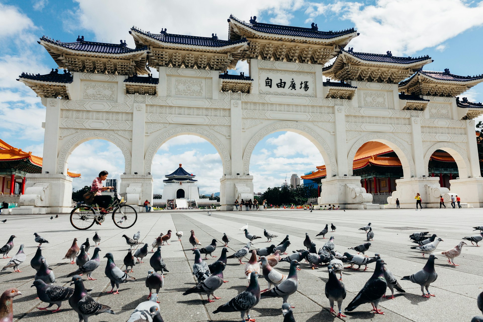 A woman cycles past the National Chiang Kai-shek Memorial Hall in Taipei on a hot sunny day with scores of pigeons walking around the square