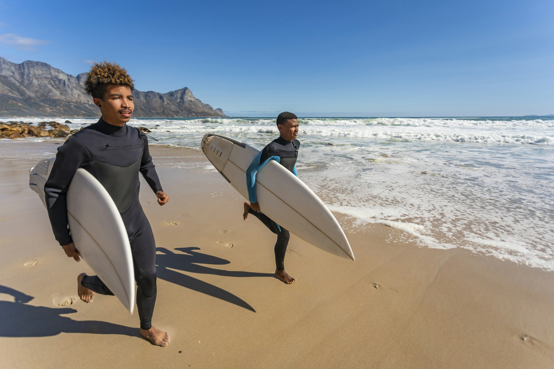 Two Black teenagers on the beach in South Africa, walking toward the water holding surfboards and carrying wetsuits