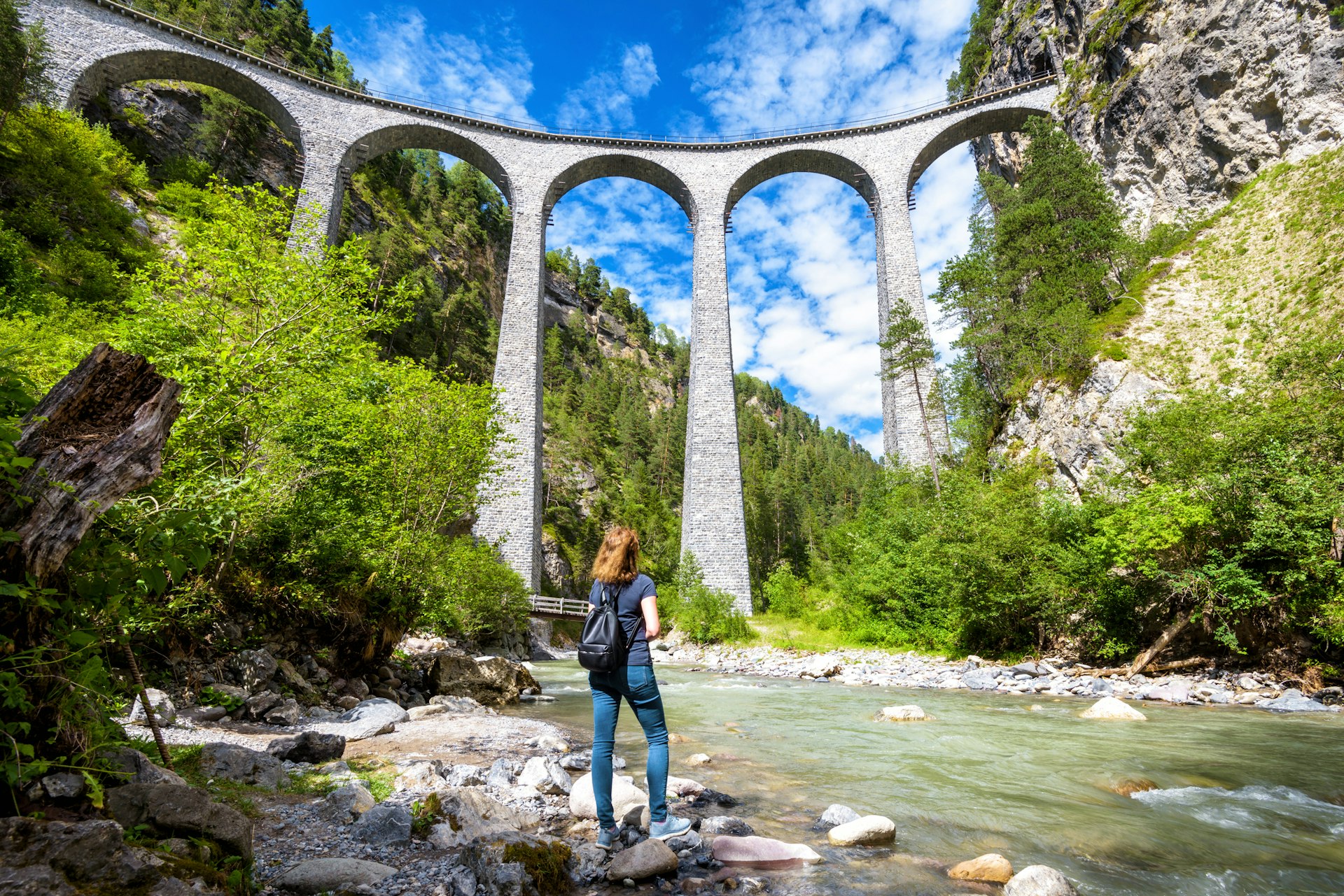 A woman stands at the edge of a river looking upwards at a multi-arched viaduct