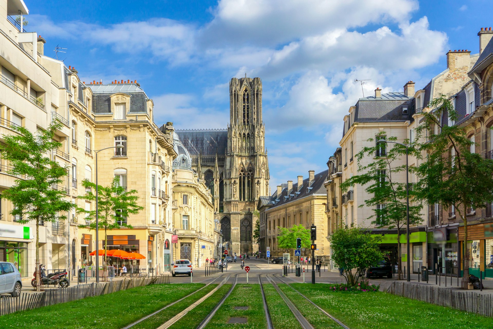 A city street lined with old buildings leading to Reims Cathedral, with green grass in the foreground