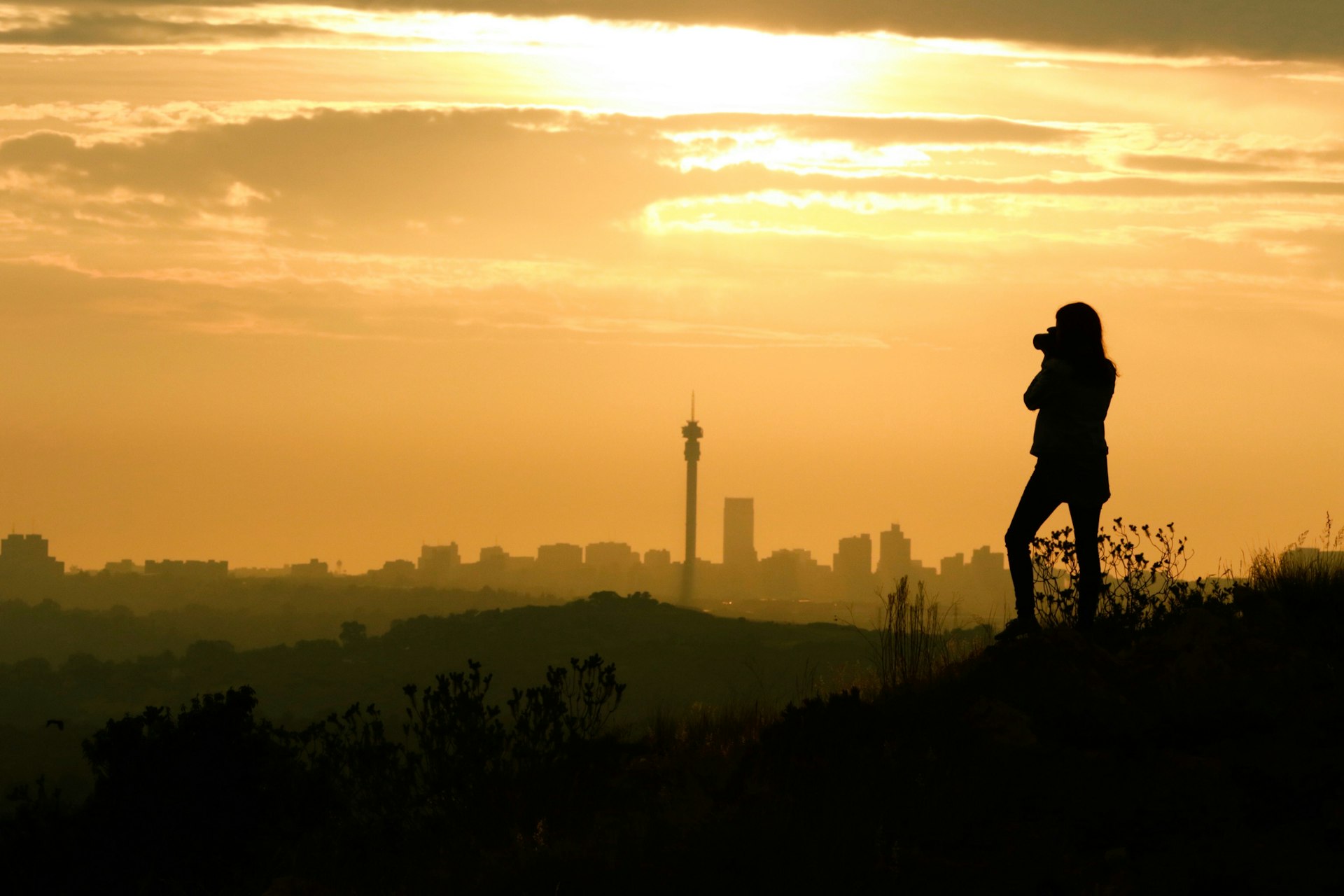 A woman in silhouette takes a photograph of the setting sun and Johannesburg city skyline in the distance