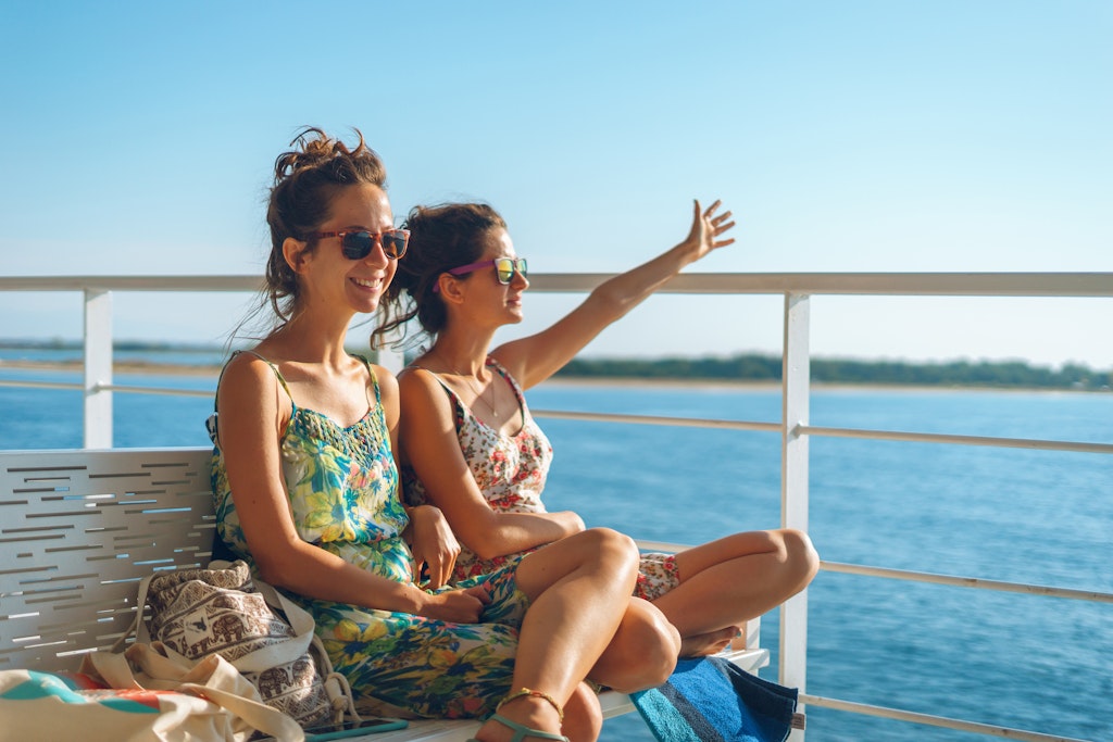Two young women girl friends or sisters sitting on the bench on the deck of the ferry boat or ship sailing to the island tourist destination on summer vacation waving to the horizon in sunny day
1176968565
Two women sit smiling on the deck of a ferry in Greece as the sun shines