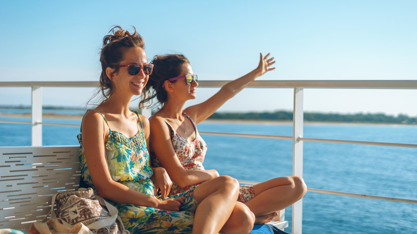 Two young women girl friends or sisters sitting on the bench on the deck of the ferry boat or ship sailing to the island tourist destination on summer vacation waving to the horizon in sunny day
1176968565
Two women sit smiling on the deck of a ferry in Greece as the sun shines