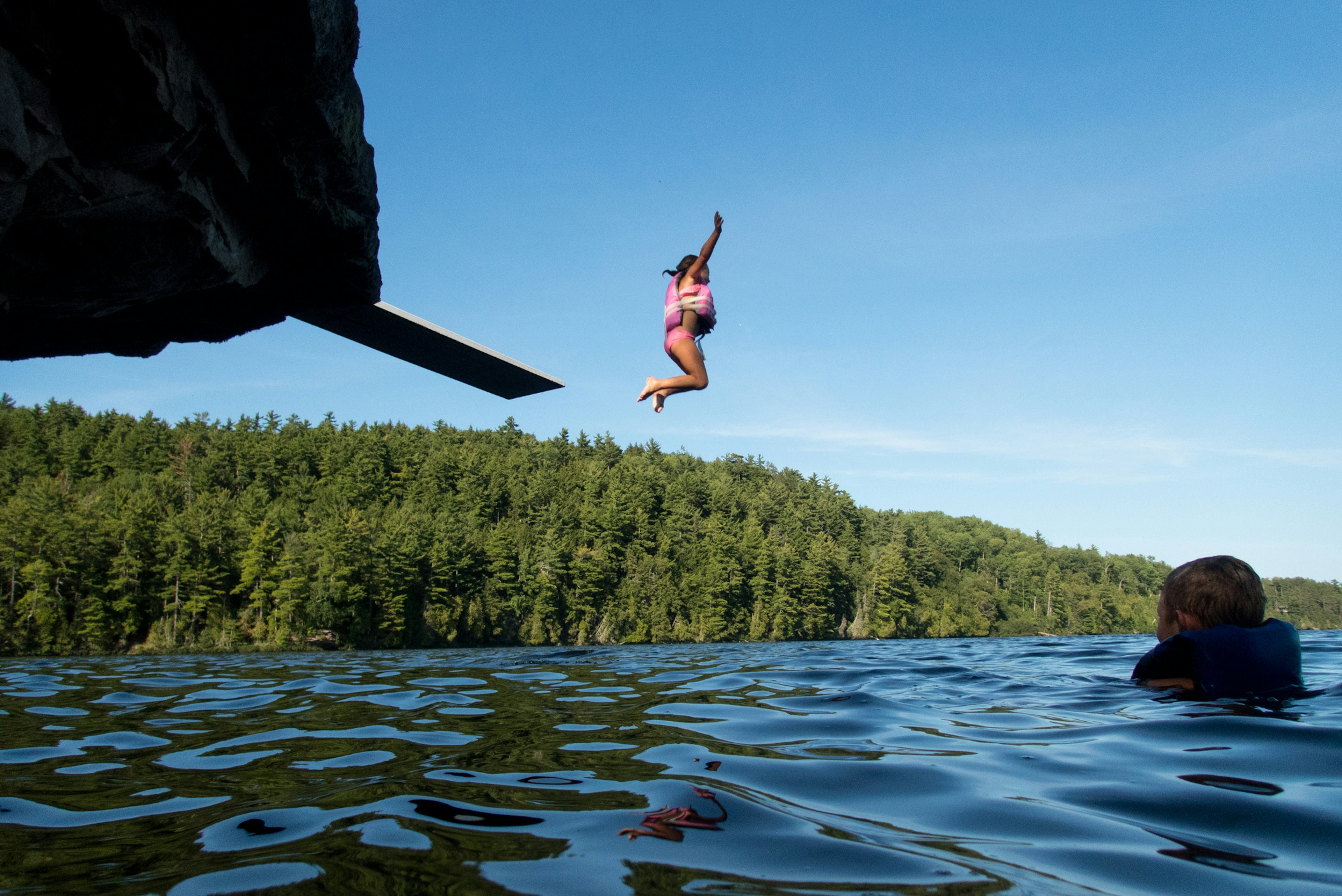 Small girl with a life jacket on jumps off a diving board into a remote lake while a boy watches from the water. Dense pine trees are visible along the shore.