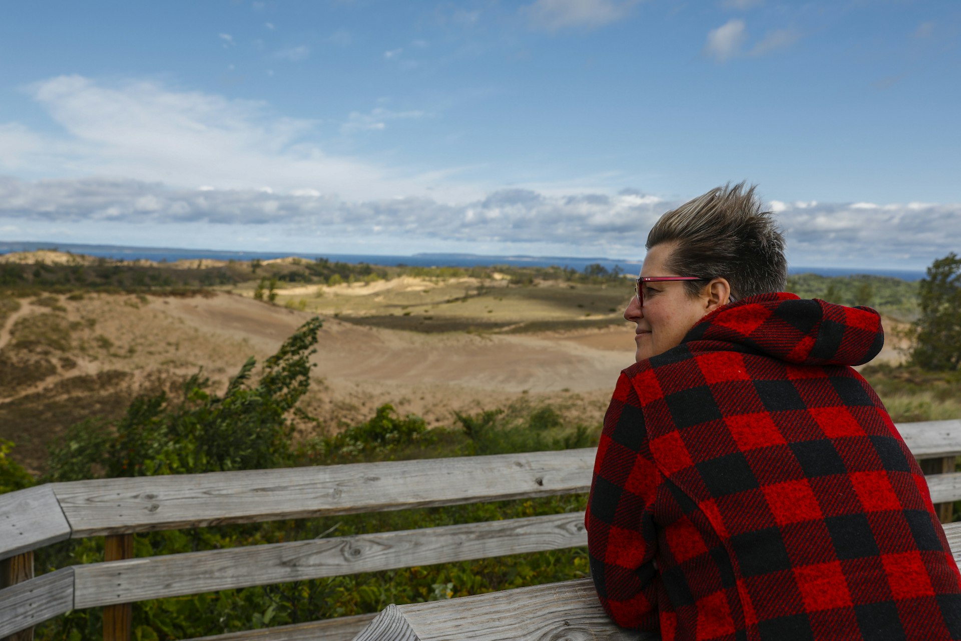 a white woman in a plaid jacket and glasses looks out across Sleeping Bear Dunes National Lakeshore with the wind whipping her short hair