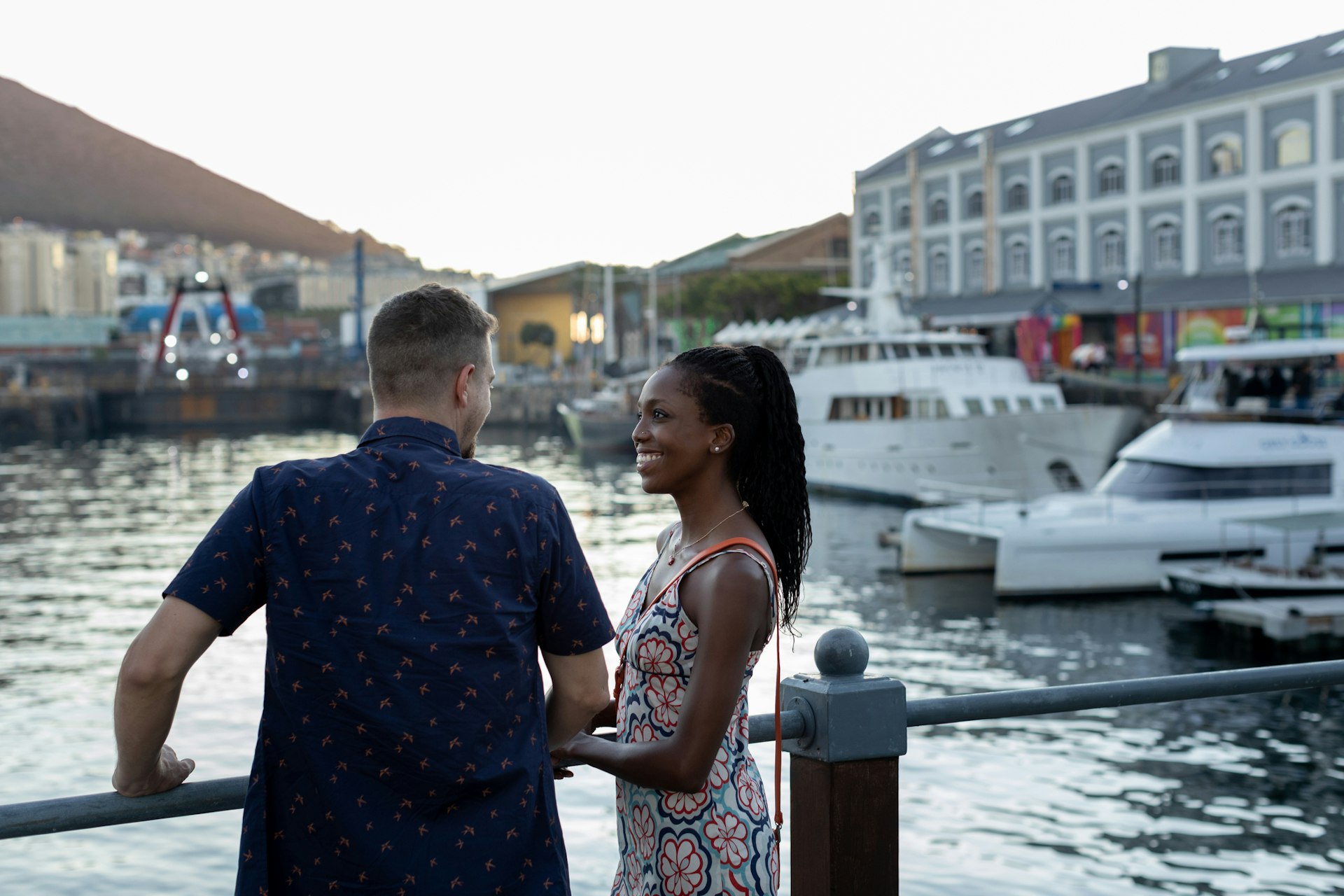 A couple stand together smiling at a waterfront where several boats are docked