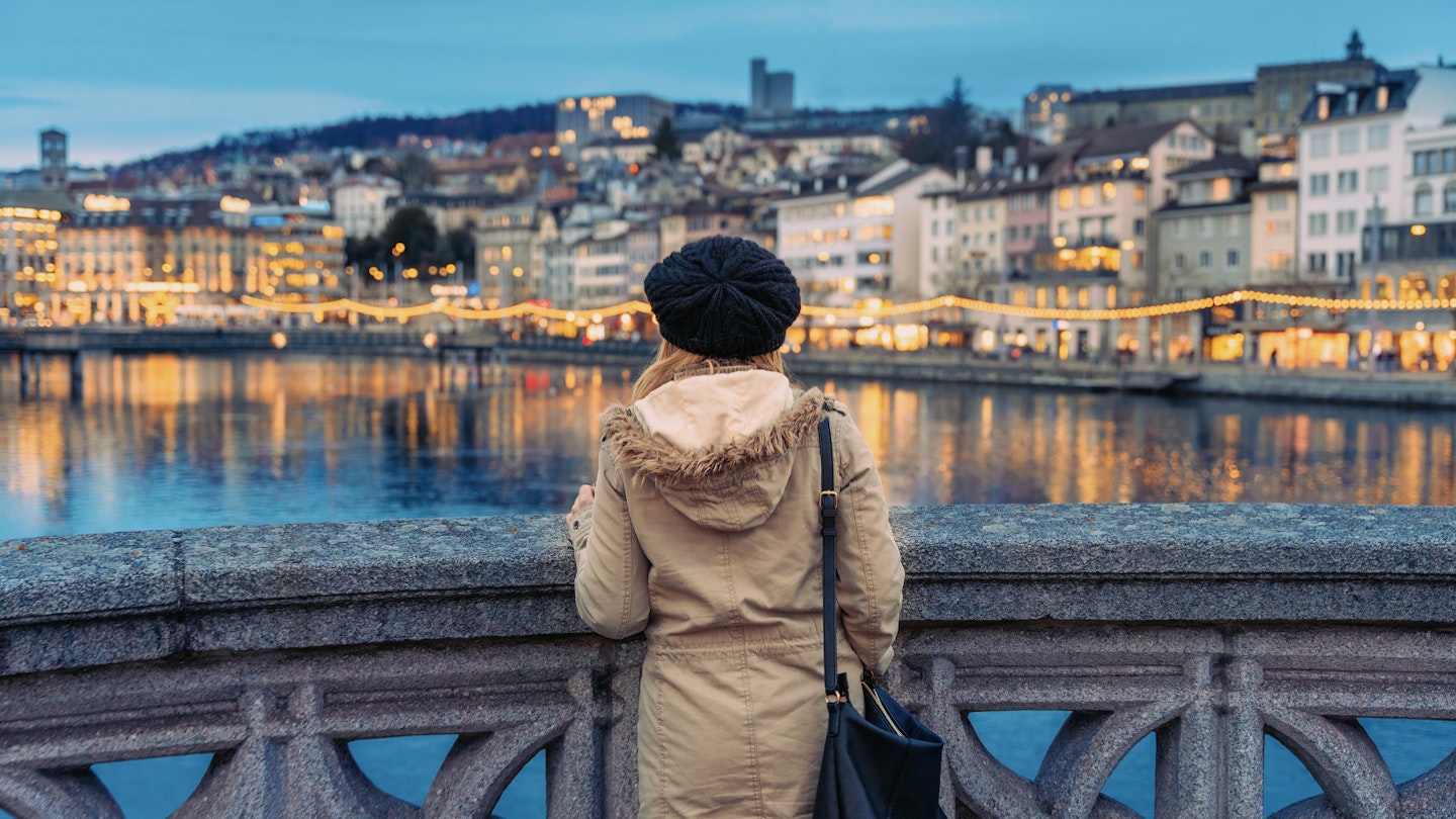Blonde girl with wool hat and heavy clothing staring at the Limmat river in central Zurich at dusk in winter
1477858415