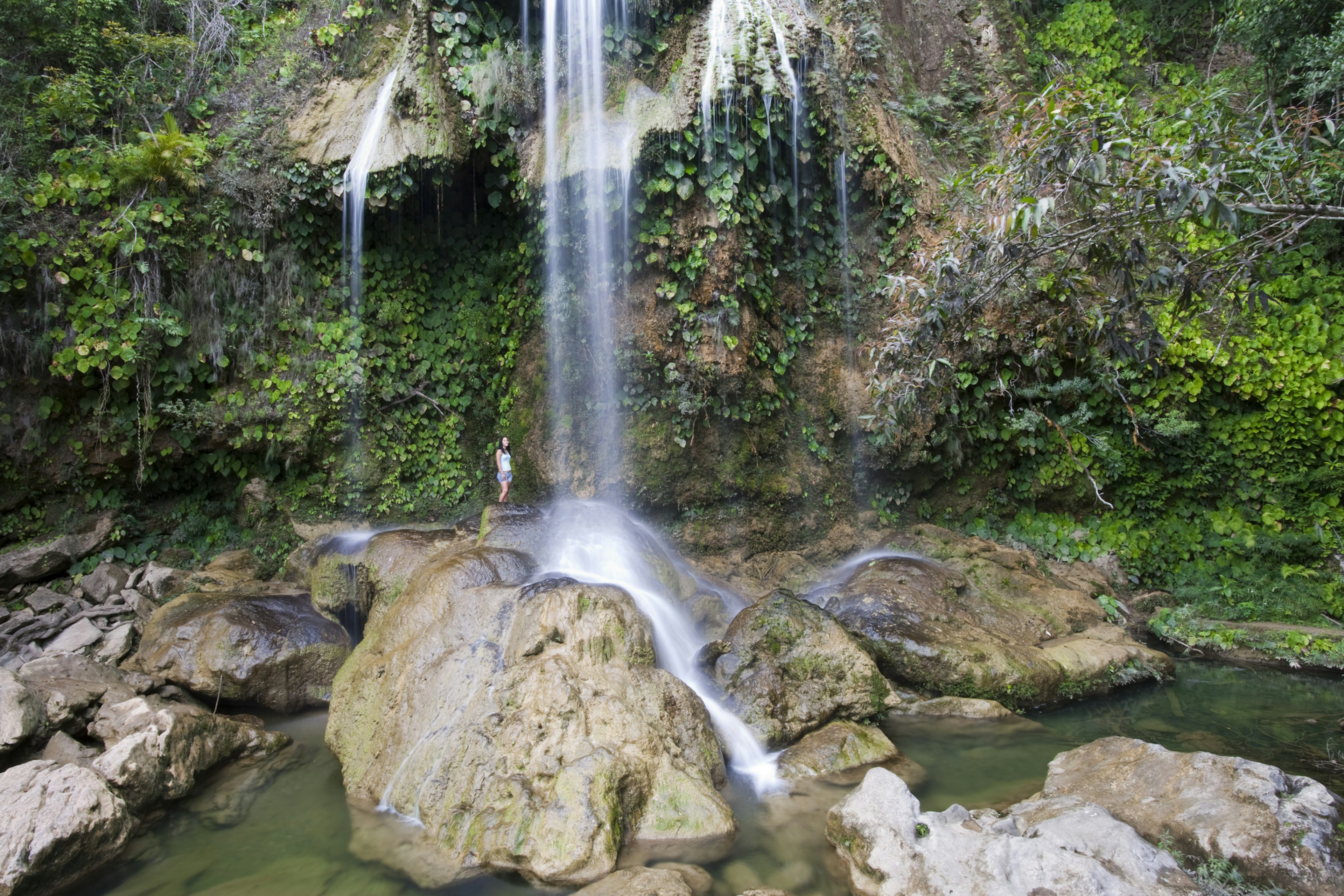 A woman stands near a cascade of water in a densely vegetated area